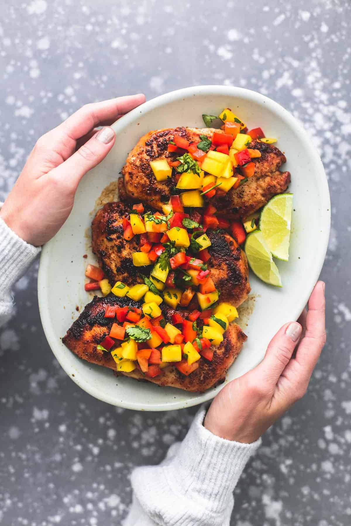 top view of a pair of hands holding a plate with mango salsa chicken and lime slices on it.