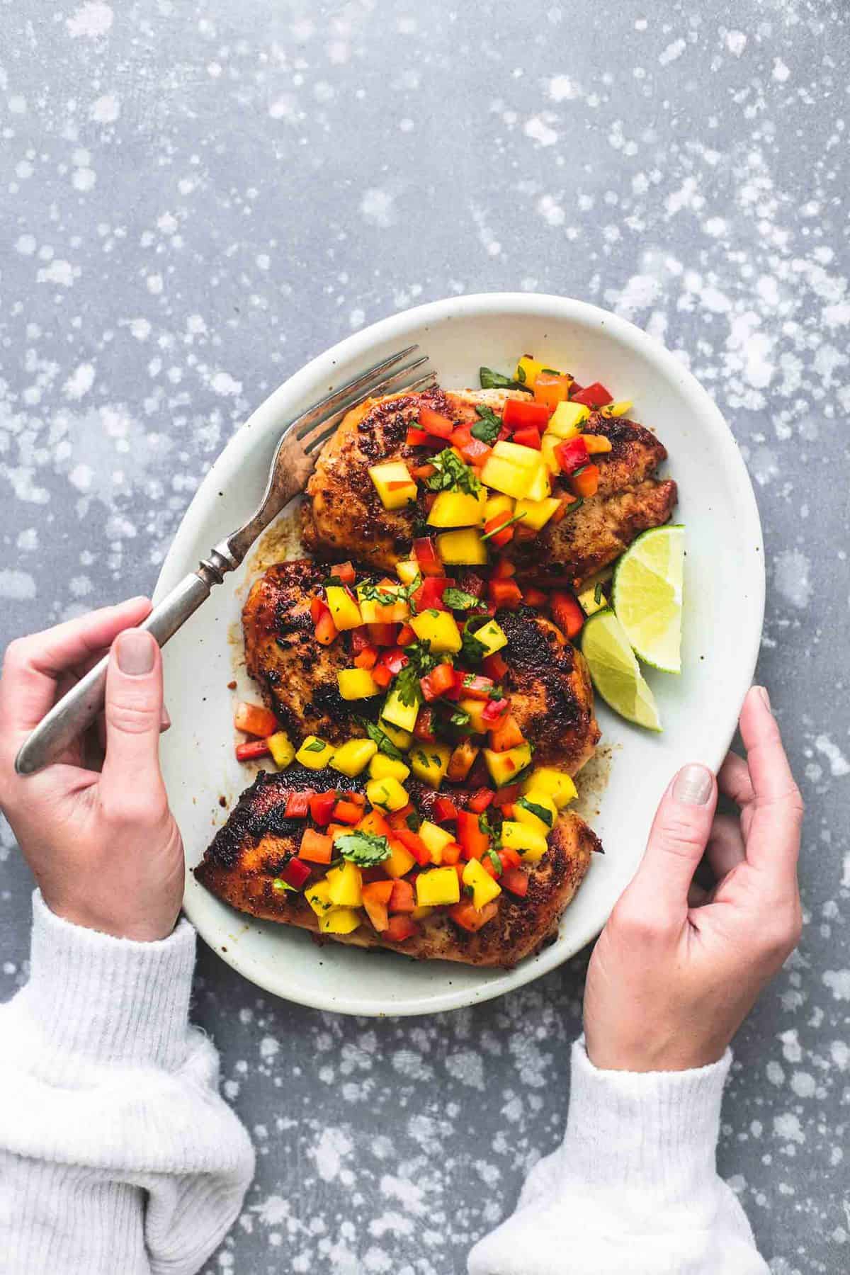 top view of one hand holding a plate with mango salsa chicken and lime slices on top with the other hand holding a fork.