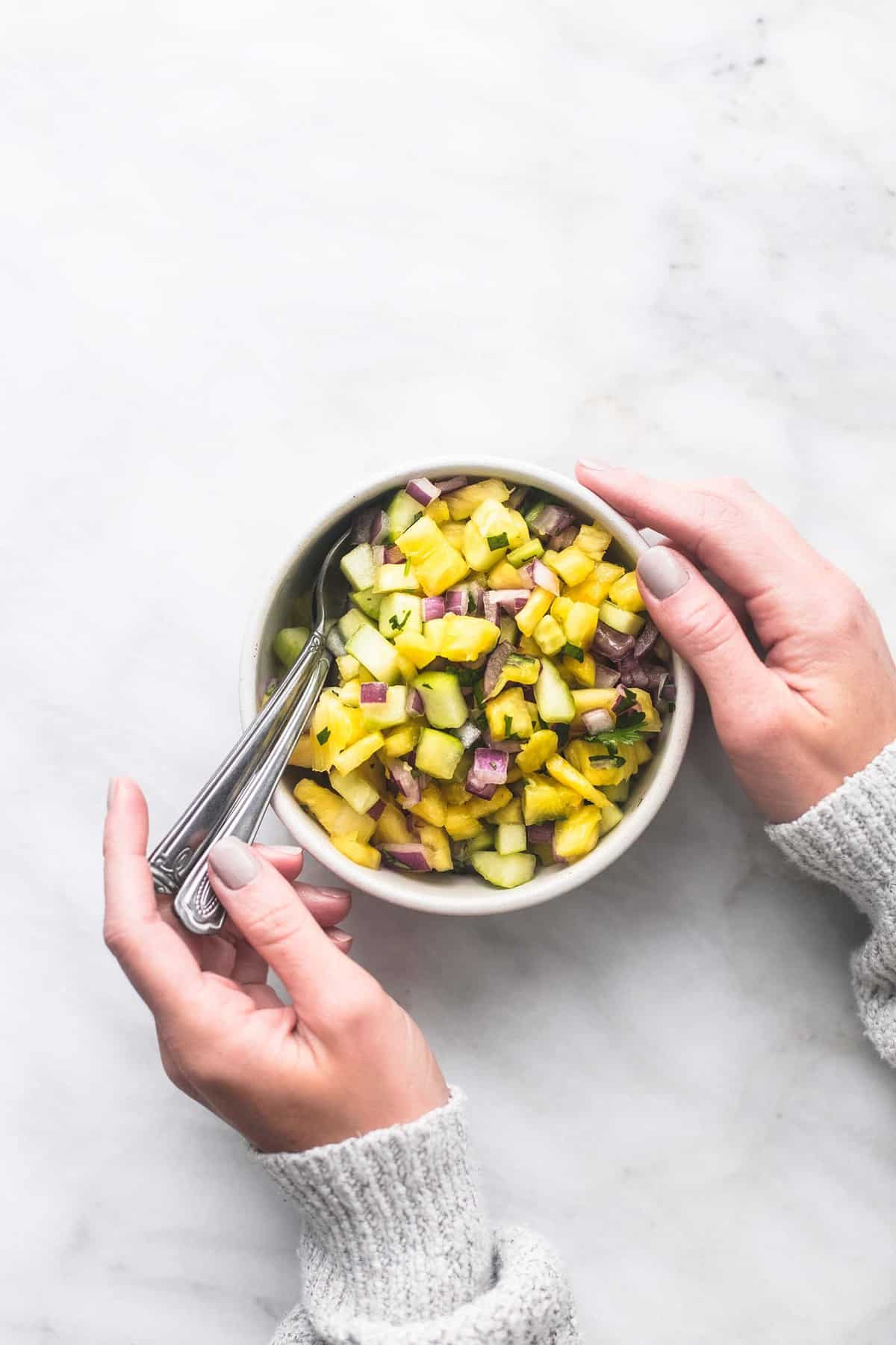 top view of pineapple salsa and spoons in a bowl with a pair of hands holding the bowl and the spoons.