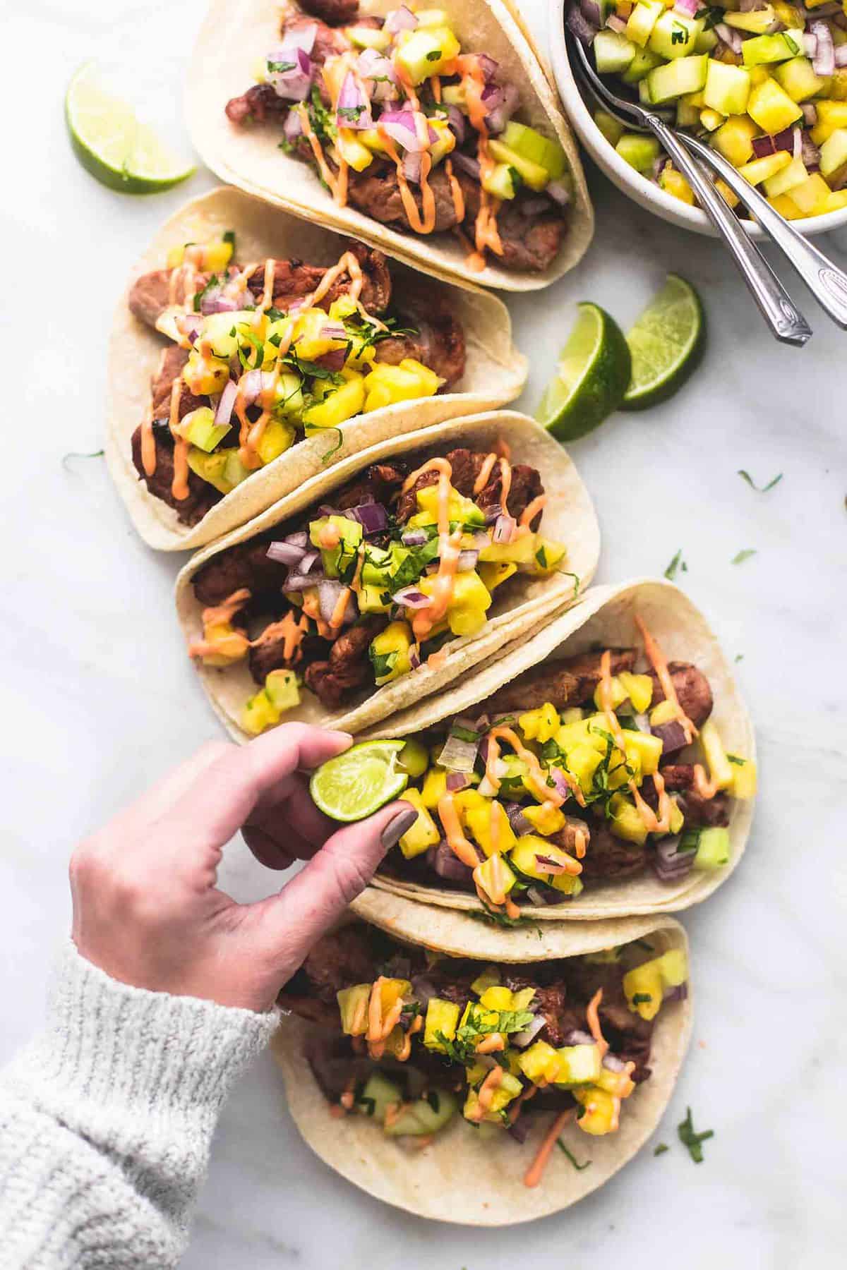 top view of pork tacos with pineapple salsa with lime slices and a bowl of salsa on the side with a hand squeezing a lemon slice over a taco.