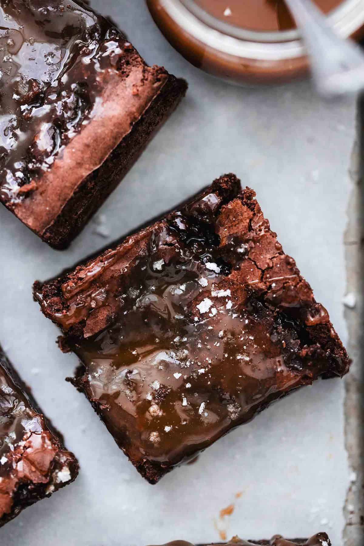 close up top view of a salted caramel brownie with more brownies and a jar of caramel with a spoon on the side.