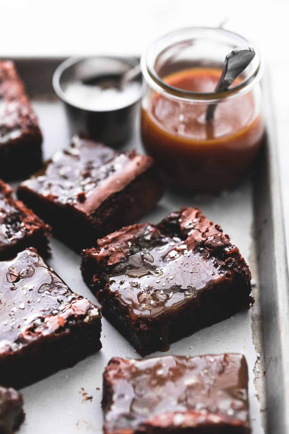salted caramel brownies, a container of salt with a spoon and a jar of caramel with a spoon in the background all on a baking sheet.