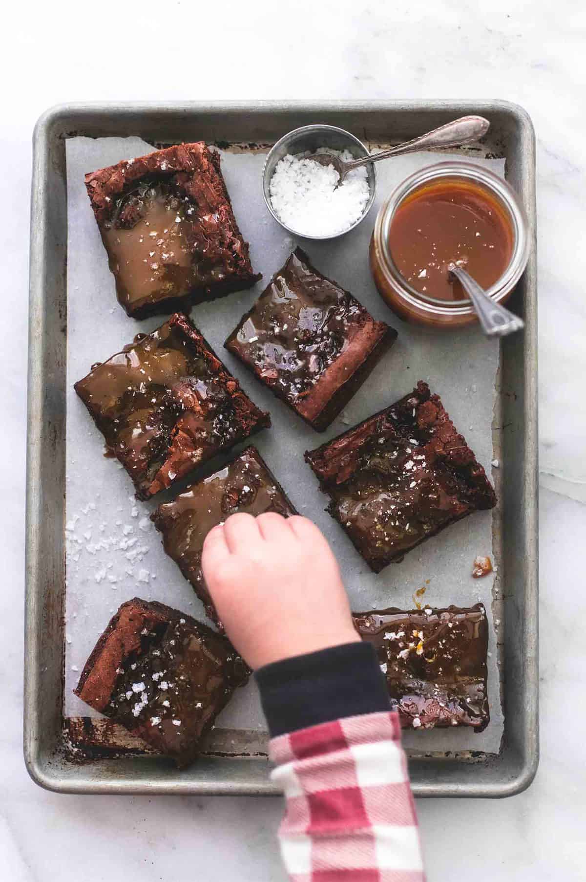 top view of a child's hand sprinkling salt over salted caramel brownies with a container of salt with a spoon and a jar of caramel with a spoon on the side all on a baking sheet.