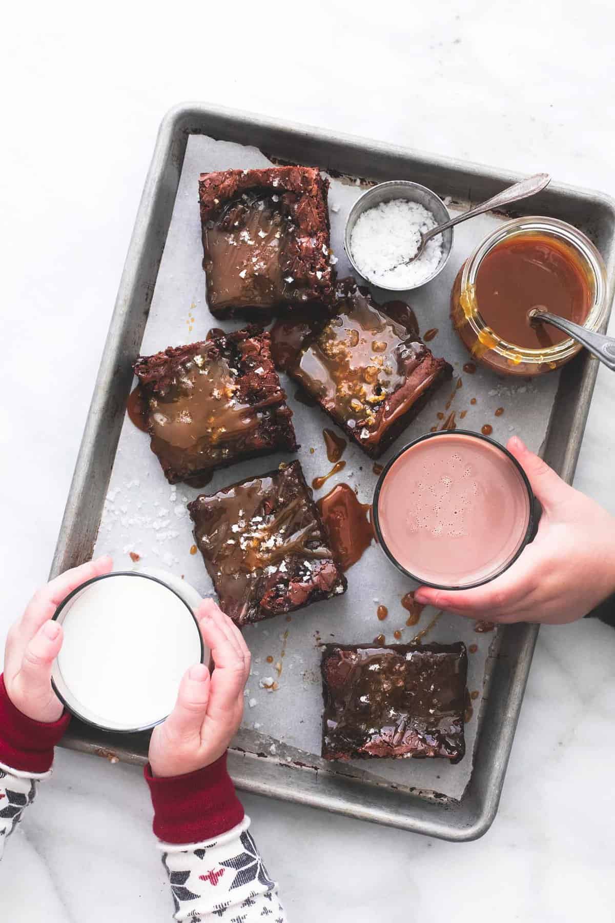 top view of salted caramel brownies, with a container of salt and a jar of caramel on the side with one child grabbing a glass of milk and another child grabbing the chocolate milk all on a baking sheet.