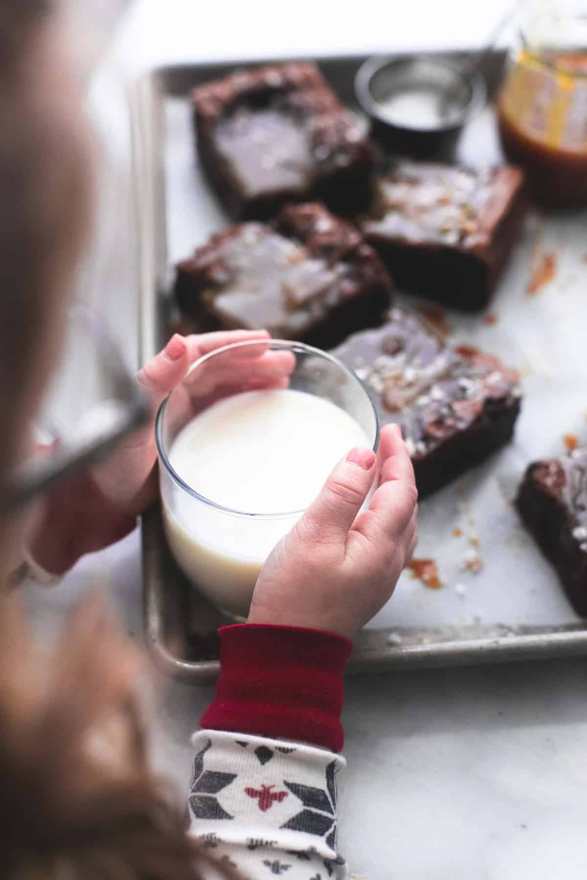 a little girl grabbing a glass of milk off of a baking pan with salted caramel brownies, a container of salt and a jar of caramel on it.