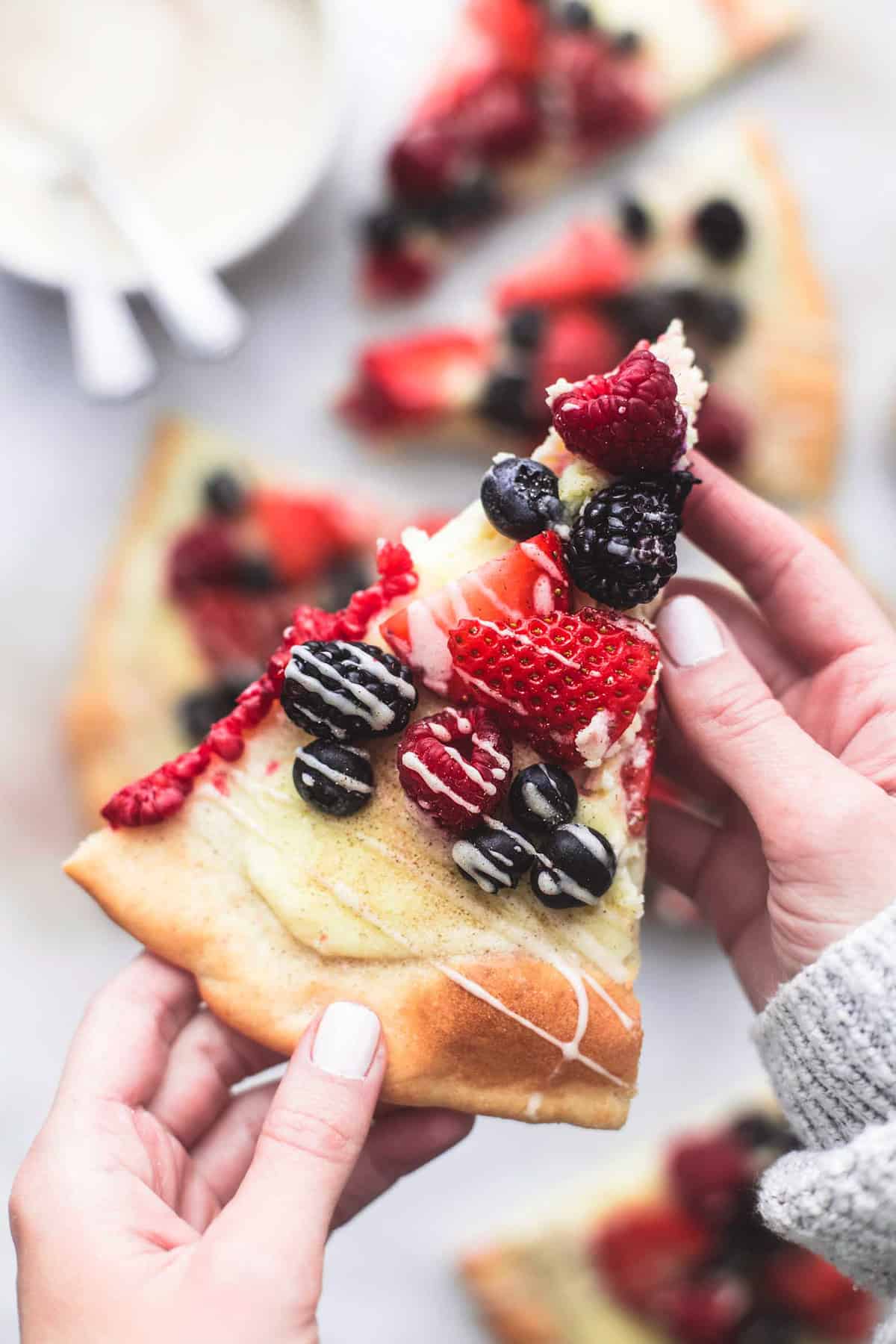 hands holding a slice of mixed berry pizza with vanilla glaze above more slices and a bowl of glaze.