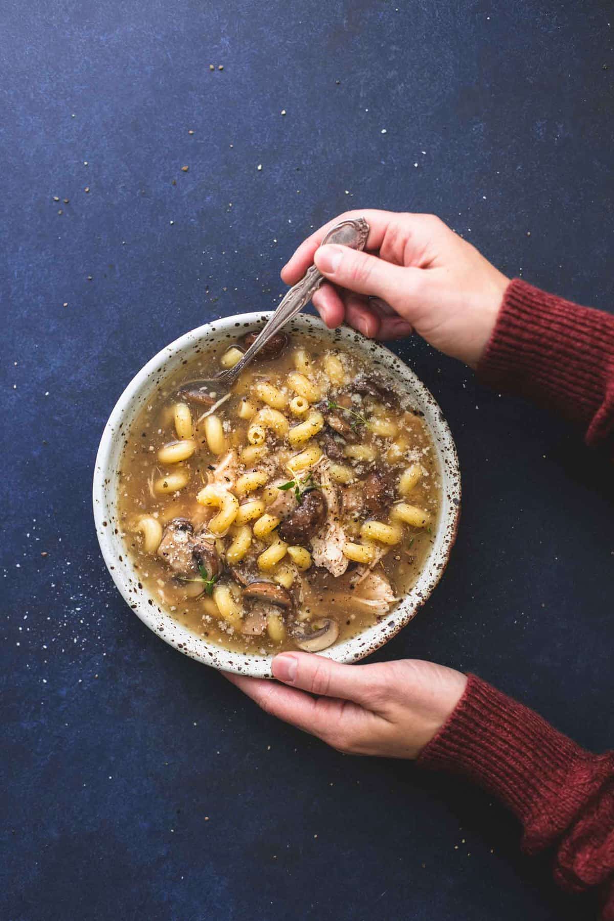 top view of a bowl of chicken marsala soup with a spoon and hands holding the bowl and spoon.