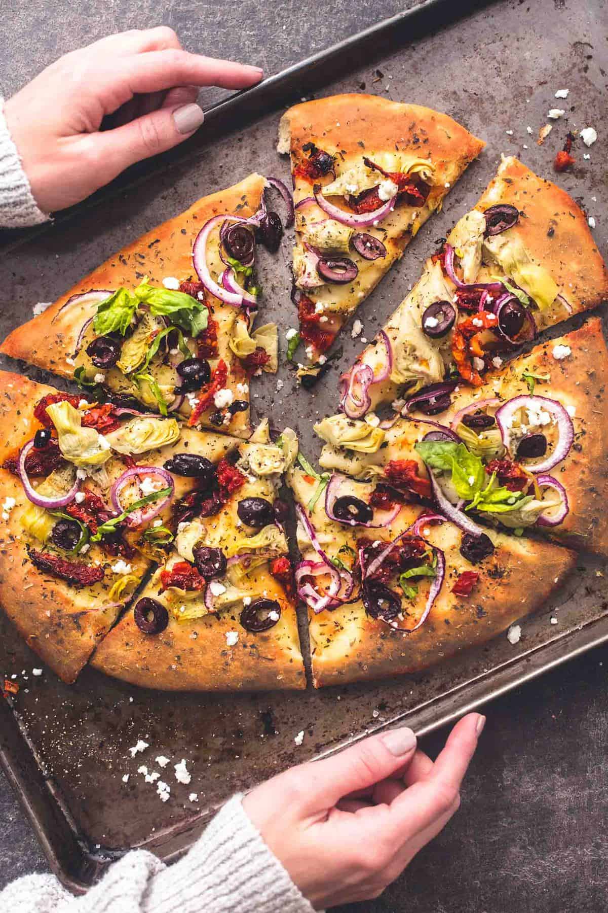 hands holding a baking sheet with Mediterranean veggie pizza slices on it.