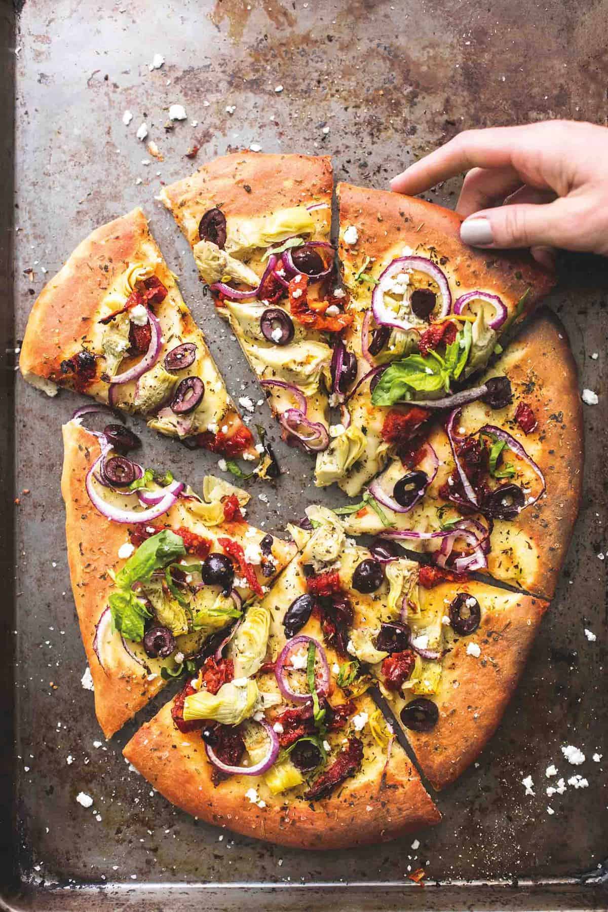 top view of Mediterranean veggie pizza slices on a baking sheet with a hand grabbing a slice.