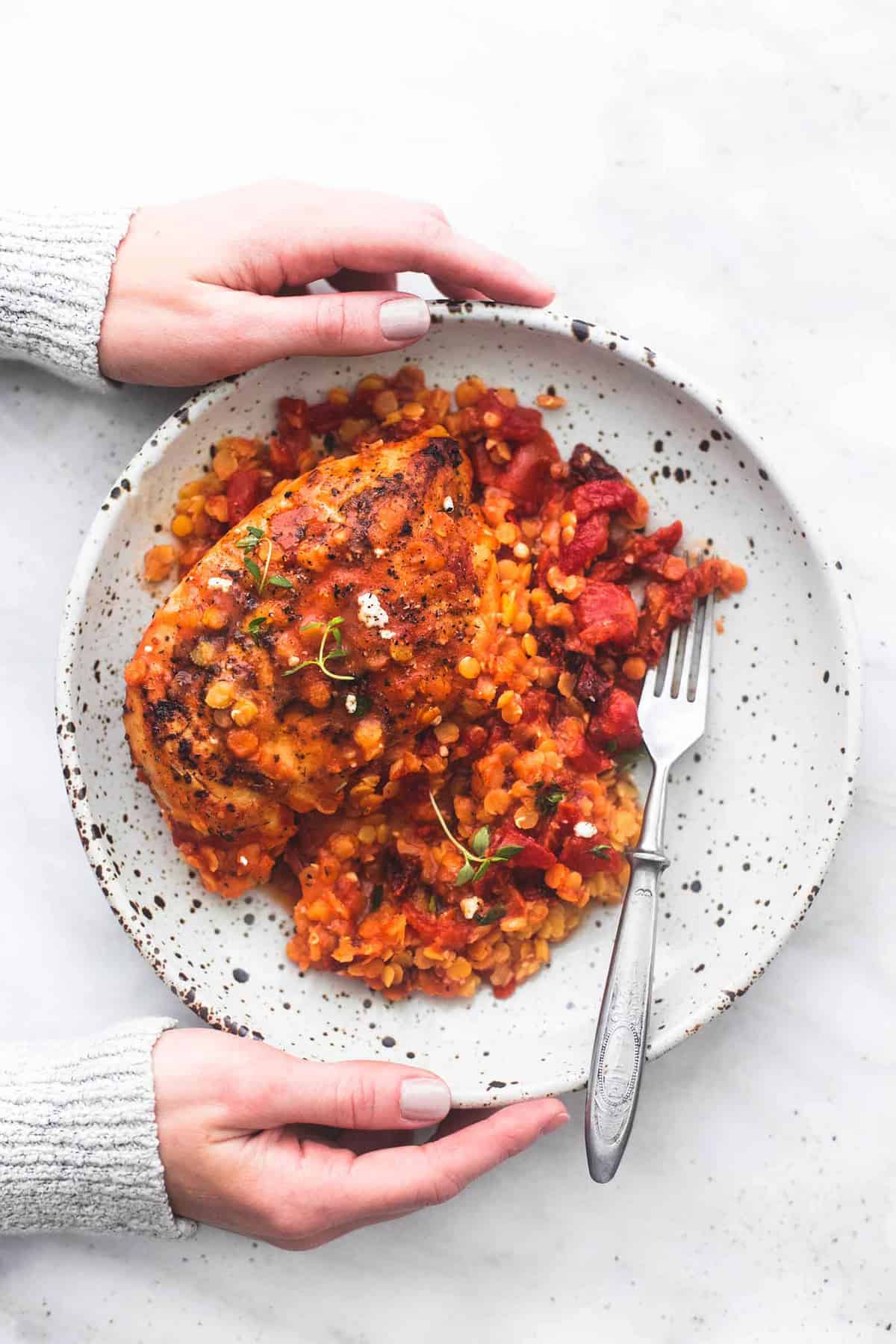 top view of a pair of hands holding a plate of Greek sun dried tomato chicken and lentils with a fork.
