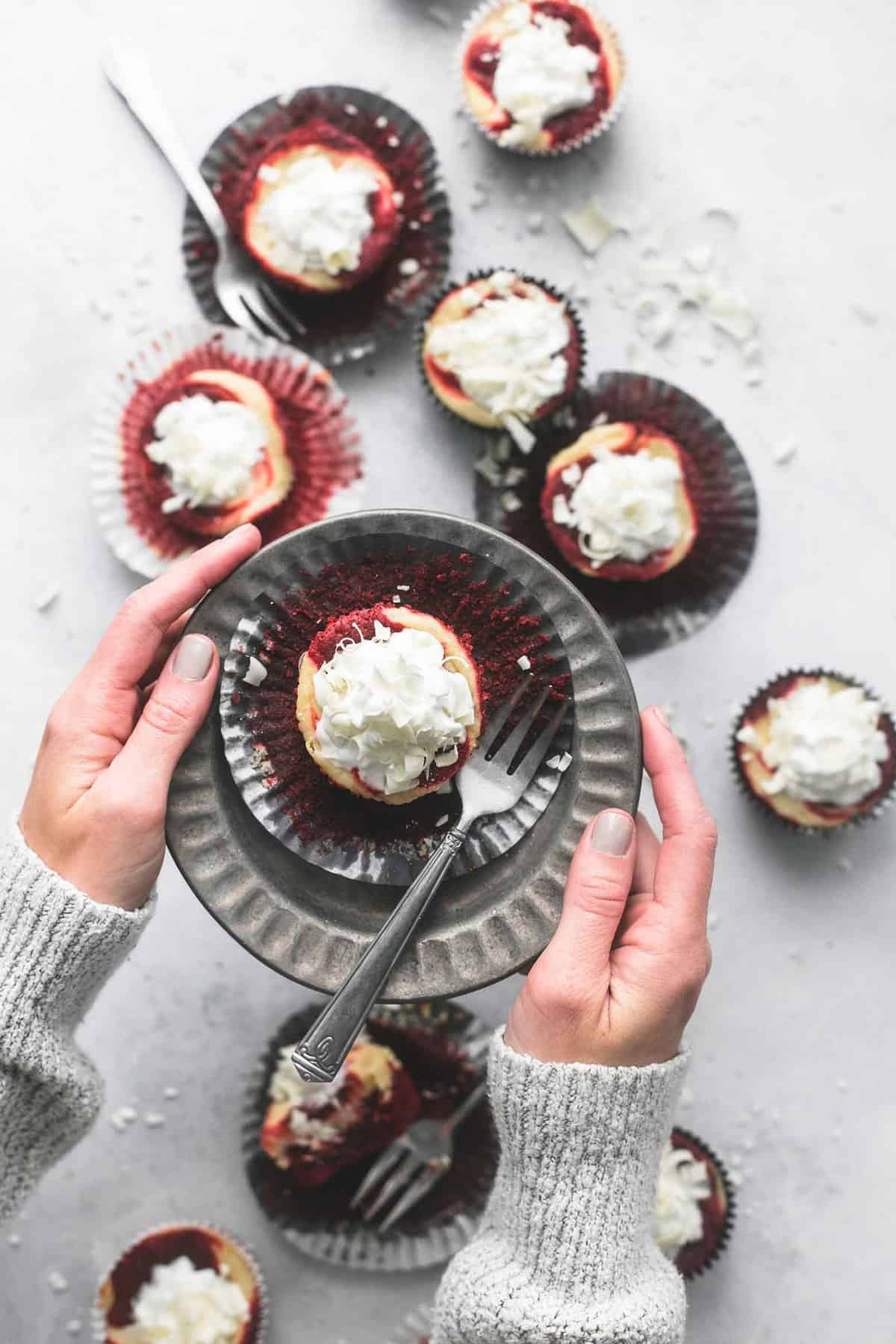 top view of hands holding a plate with a red velvet cheesecake bites and a fork on top above more cheesecake bites.