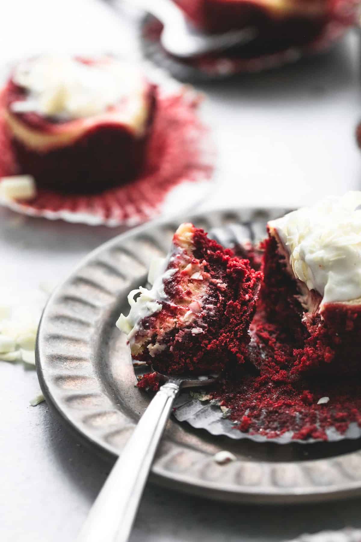 a bite of a red velvet cheesecake bite on a fork next to the rest of the cheesecake bite on a plate with more cheesecake bites in the background.