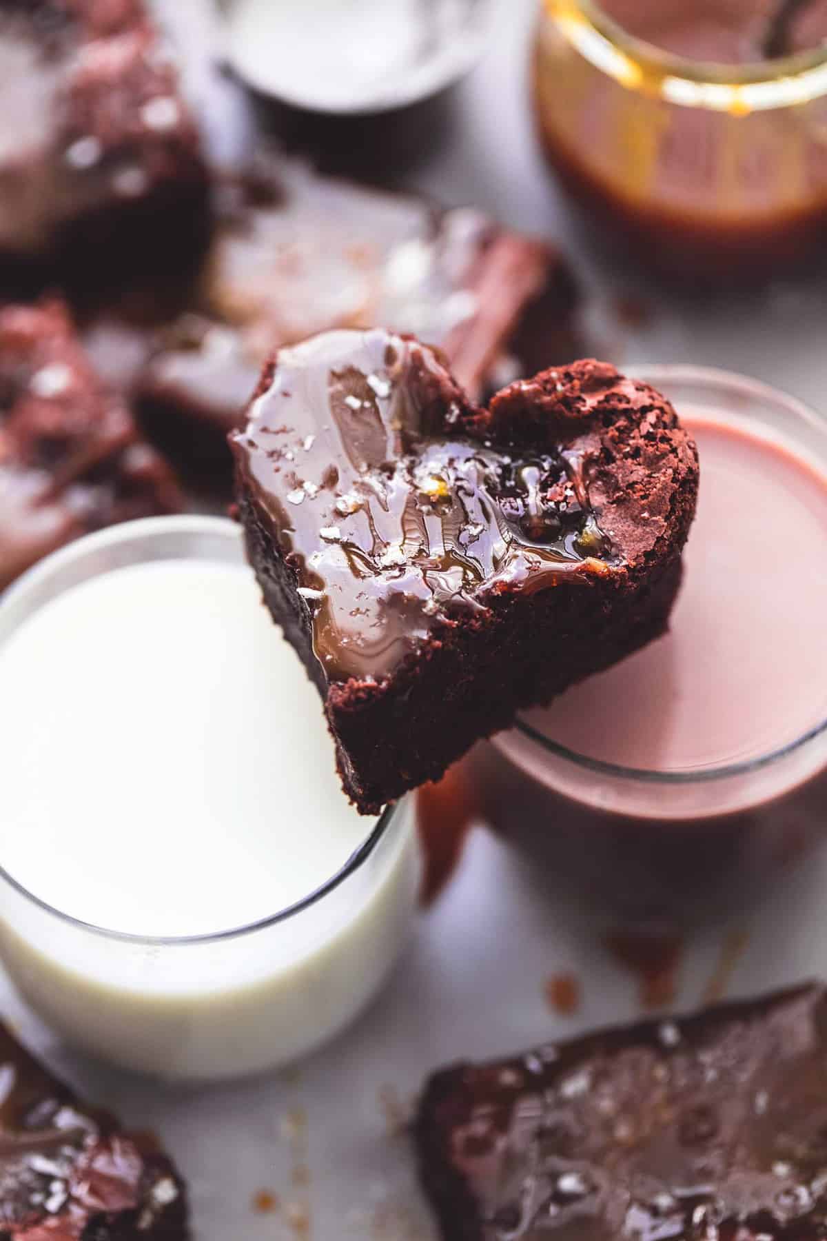 a salted caramel brownie sitting on top of two glasses of milk and chocolate milk with more brownies and a jar of caramel on the side.