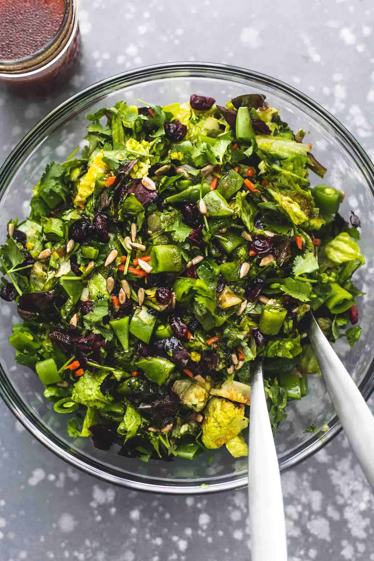 top view of chopped Asian salad with two serving spoons in a glass bowl with a jar of teriyaki dressing on the side.