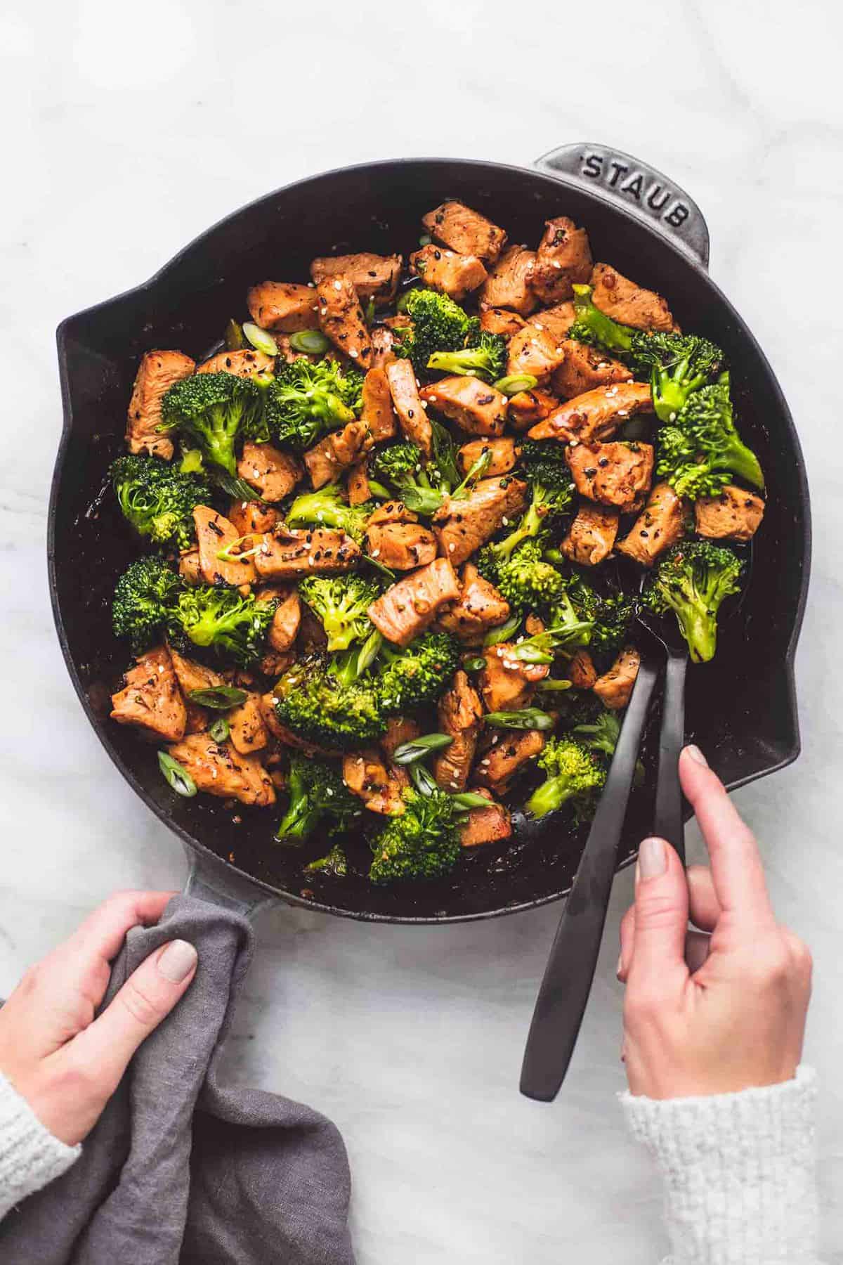 top view of chicken and broccoli stir fry in a pan with a hand on the handle and another hand on one of the serving swoons in the pan.