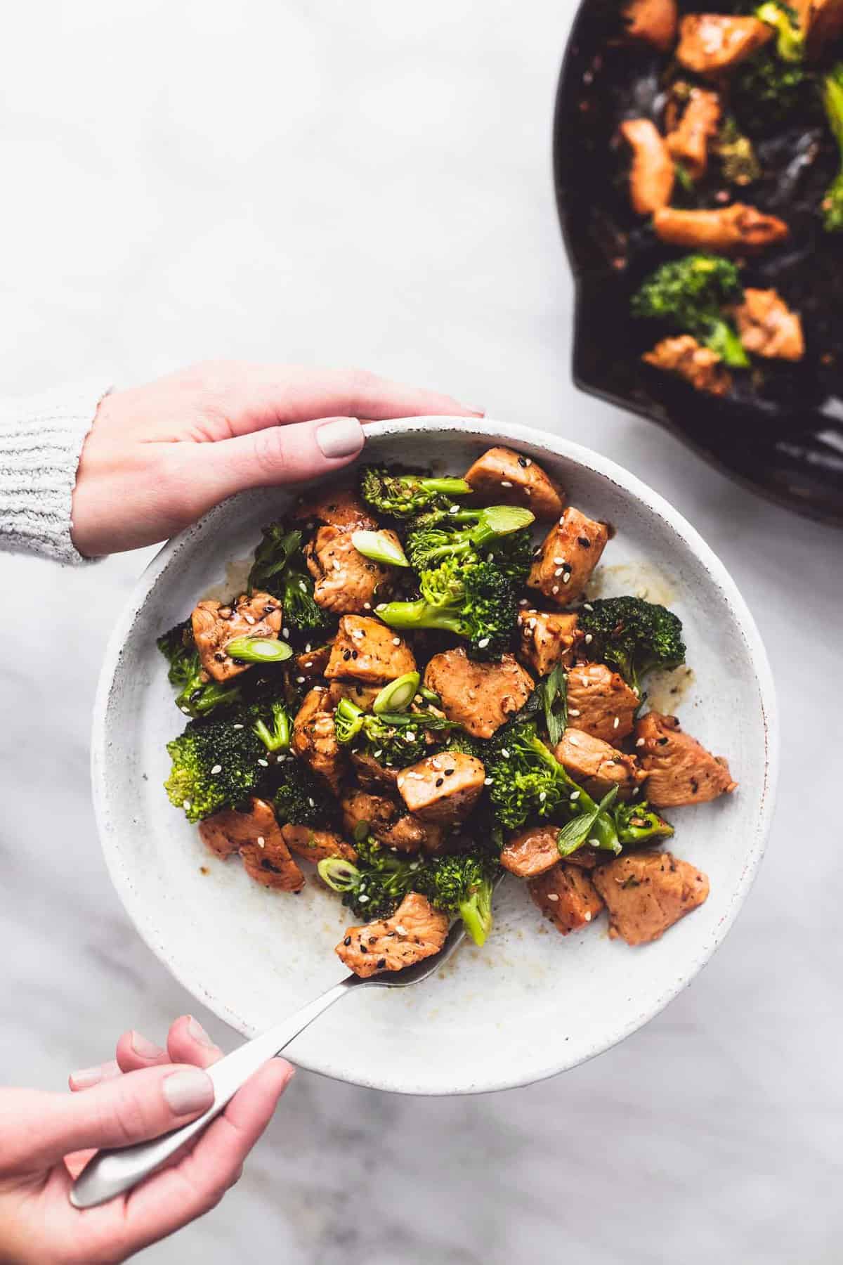 top view of chicken and broccoli stir fry with a hand holding a plate and grabbing a bite with a fork with the other hand.