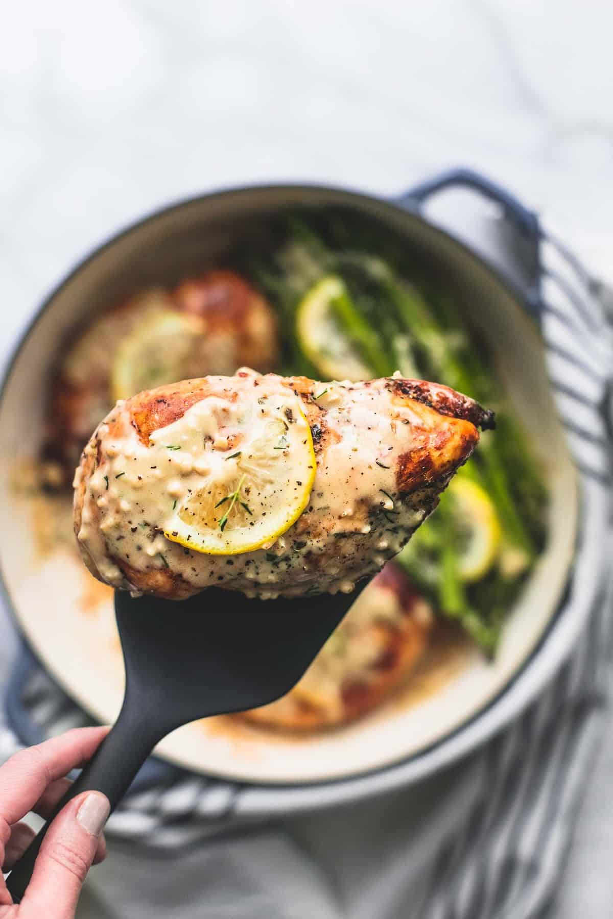 a piece of creamy lemon chicken on a spatula being held up above a pan of more chicken and asparagus.