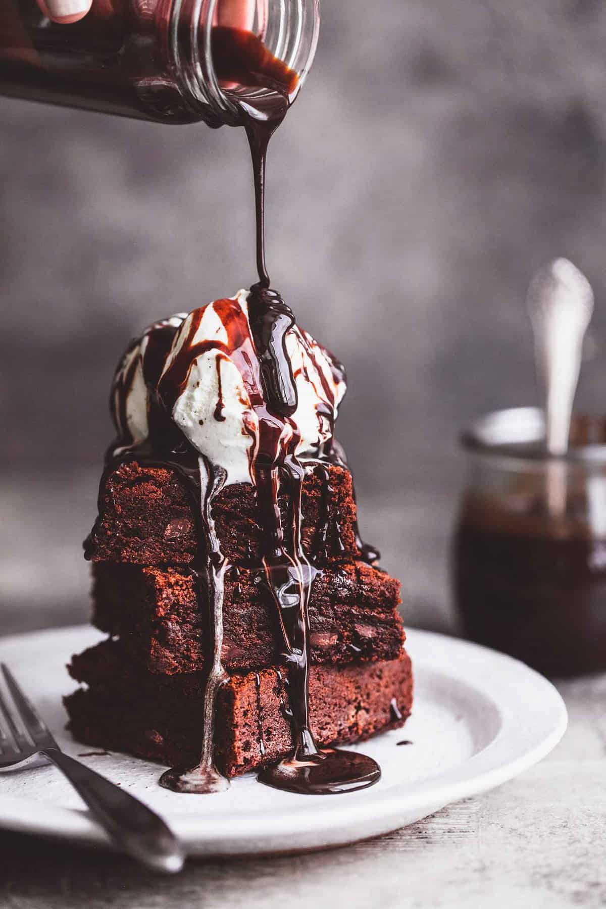 a jar of hot fudge being poured on top of a stack of brownies with a scoop of ice cream on the top with a fork on the side on a plate.