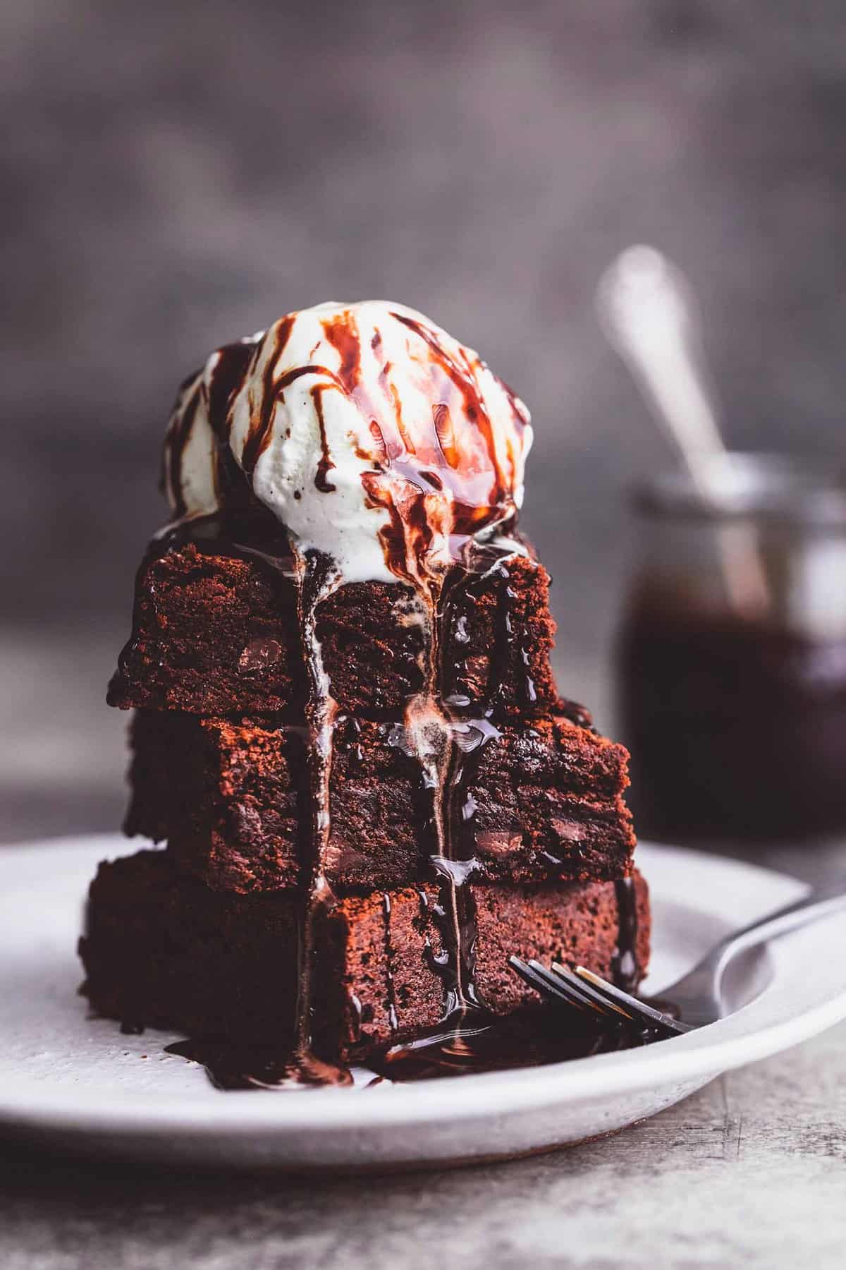 brownies stacked on a plate with a scoop of ice cream and hot fudge on top with a fork on the side and a jar of hot fudge with a spoon in the background.