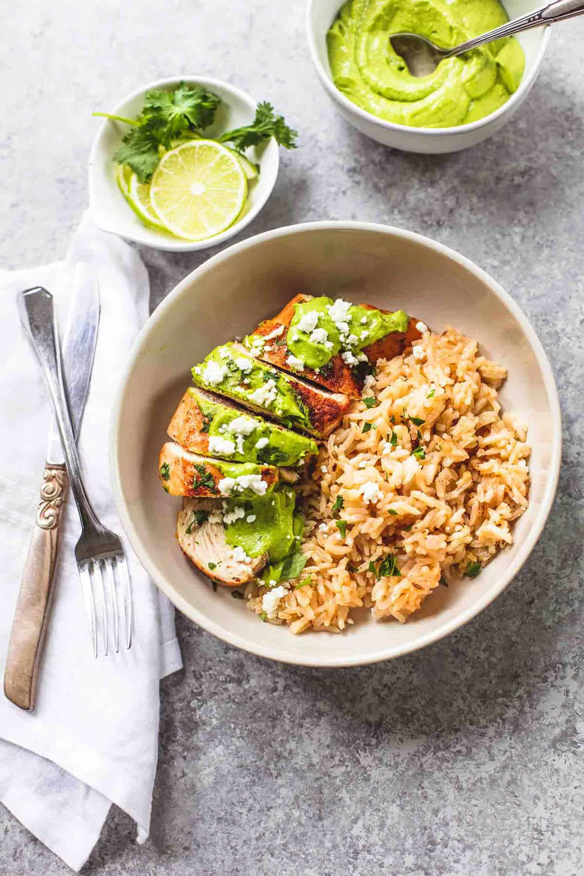a bowl of one pan creamy cilantro lime chicken and rice with a fork and knife overlapping on a cloth napkin, a bowl of lime wheels and cilantro and a bowl of sauce on the side.