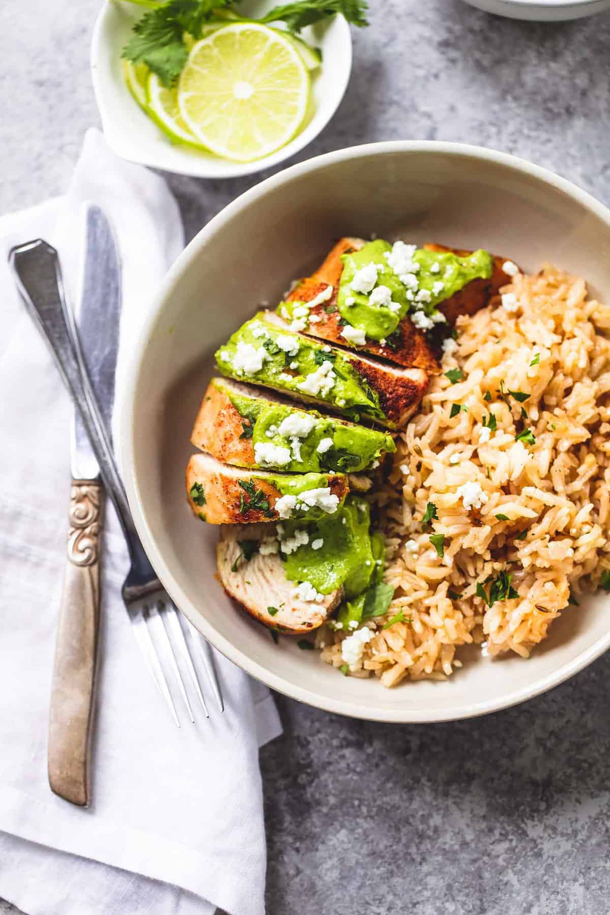 top view of a bowl of one pan creamy cilantro lime chicken and rice with a knife and fork overlapping on a cloth napkin and a bowl of lime wheels on the side.