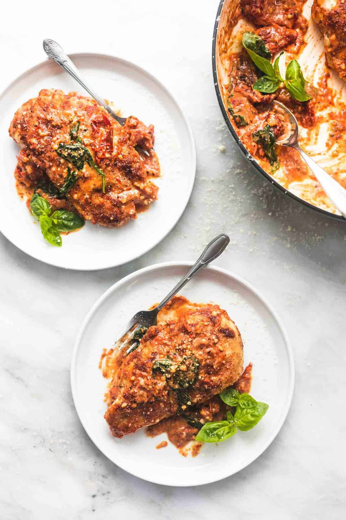 top view of two plates with creamy Tuscan parmesan chicken and forks on them next to a pan of more chicken.