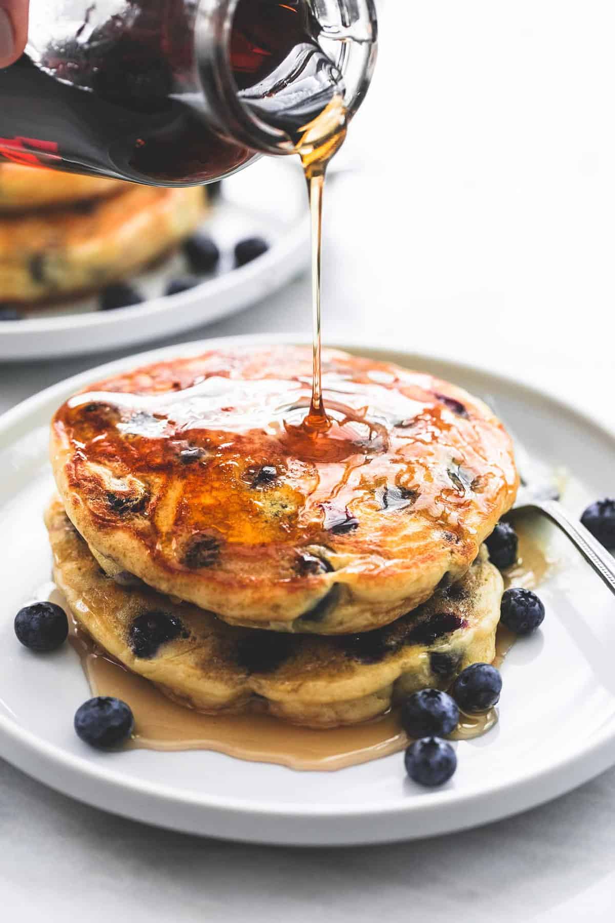 a jar of syrup being poured on top of a stack of blueberry pancakes with blueberries and a fork on the side all on a plate.