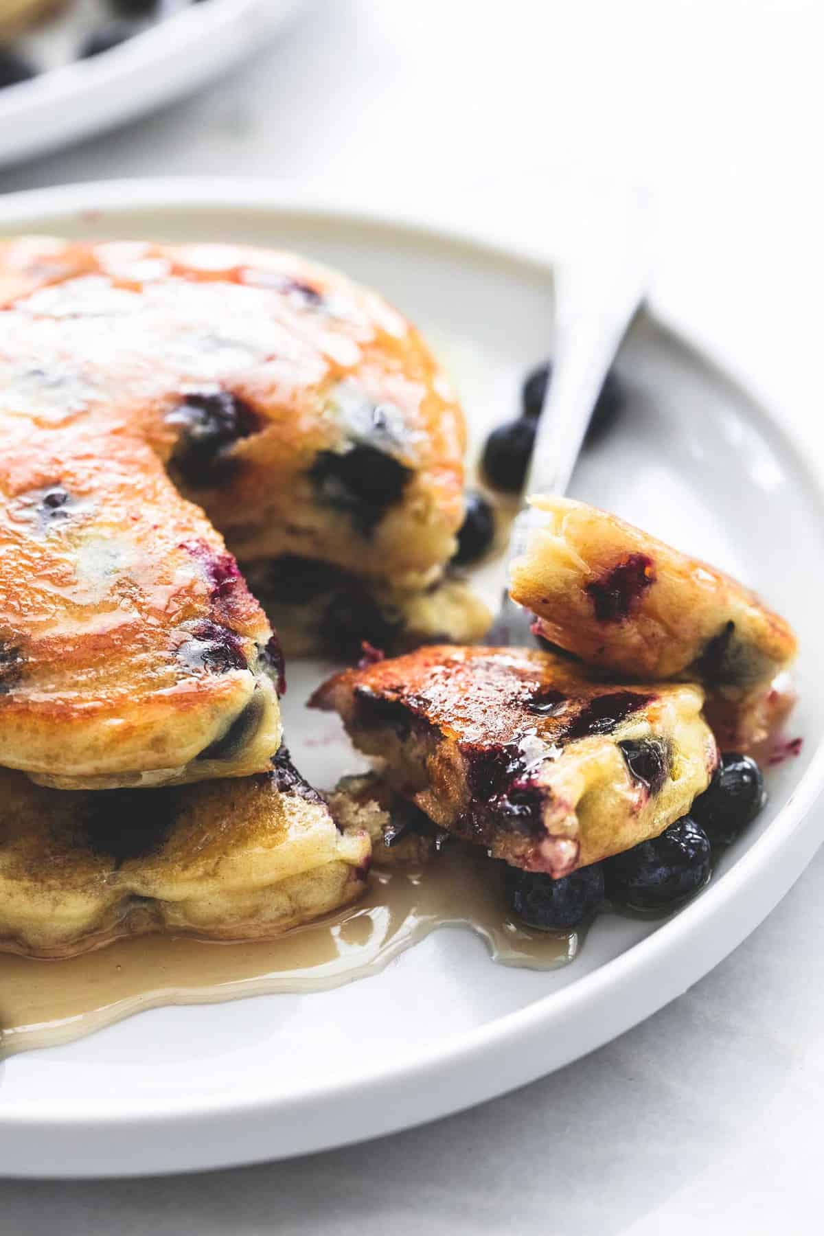 close up of a stack of blueberry pancakes with syrup with a bite on a fork on a plate.
