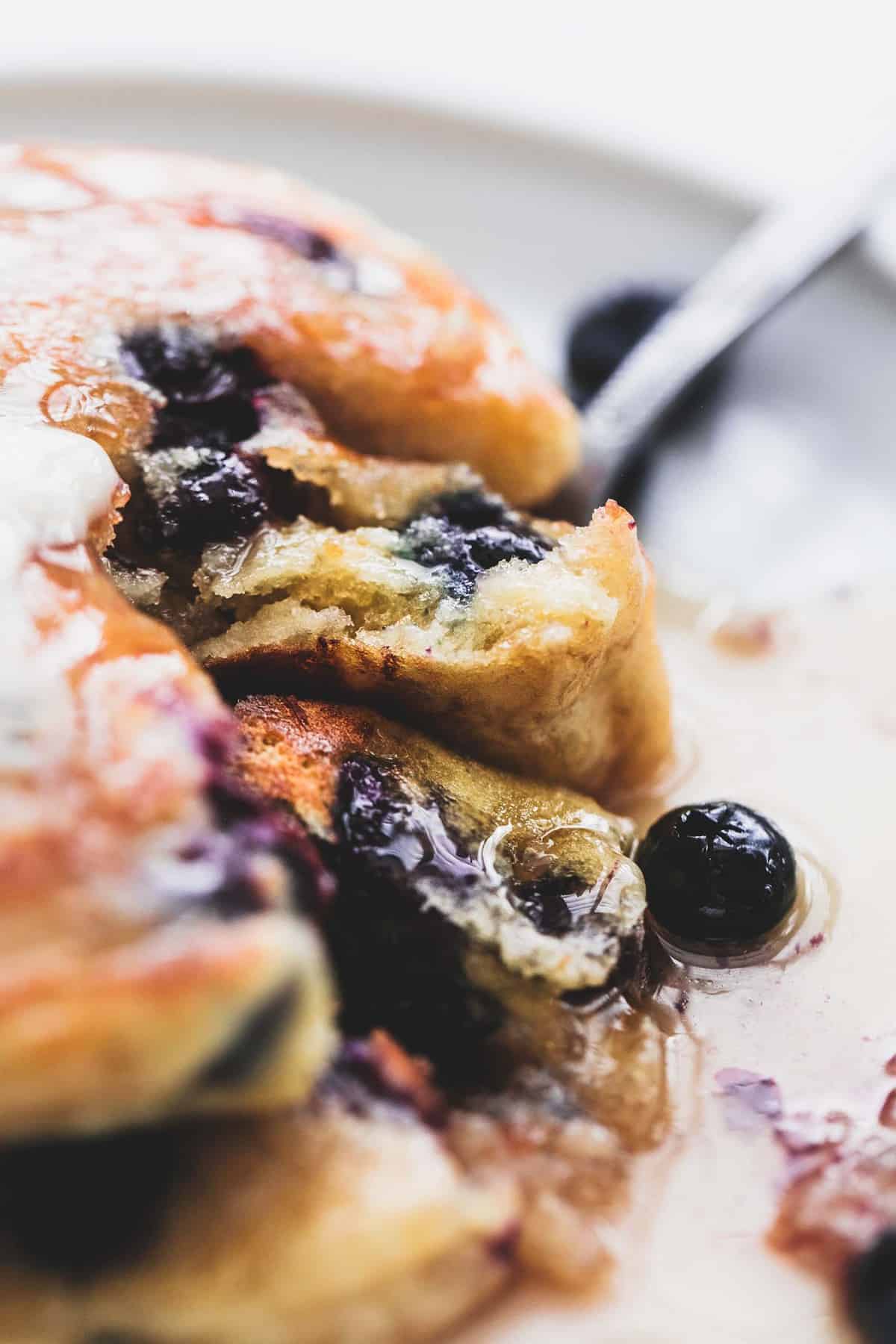 close up of blueberry pancakes topped with syrup on a plate with a fork separating a bite form the rest of the stack.