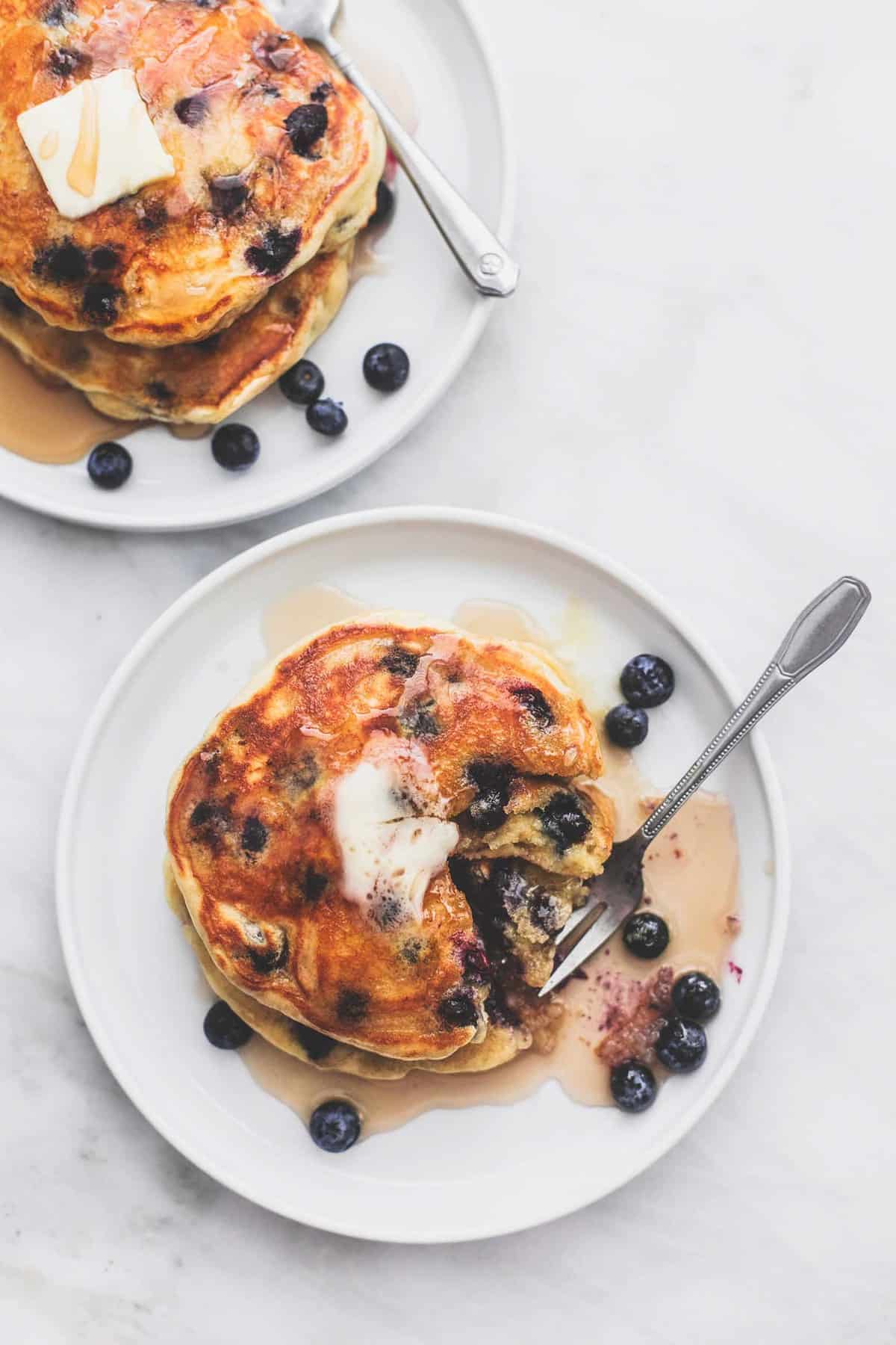 top view of a stack of blueberry pancakes with a fork underneath a bite with another plate of pancakes on the side.