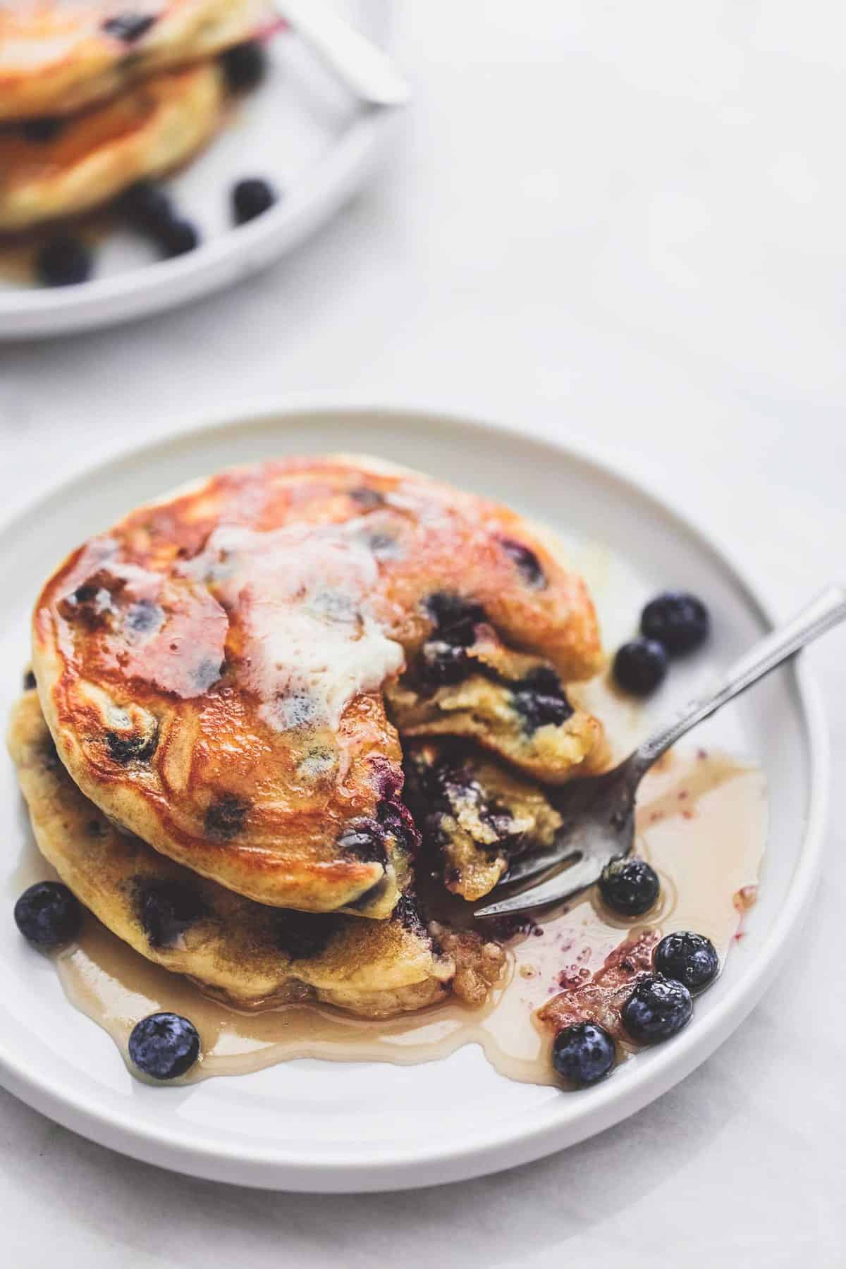 a stack of blueberry pancakes topped with syrup on a plate with a bite underneath a fork.