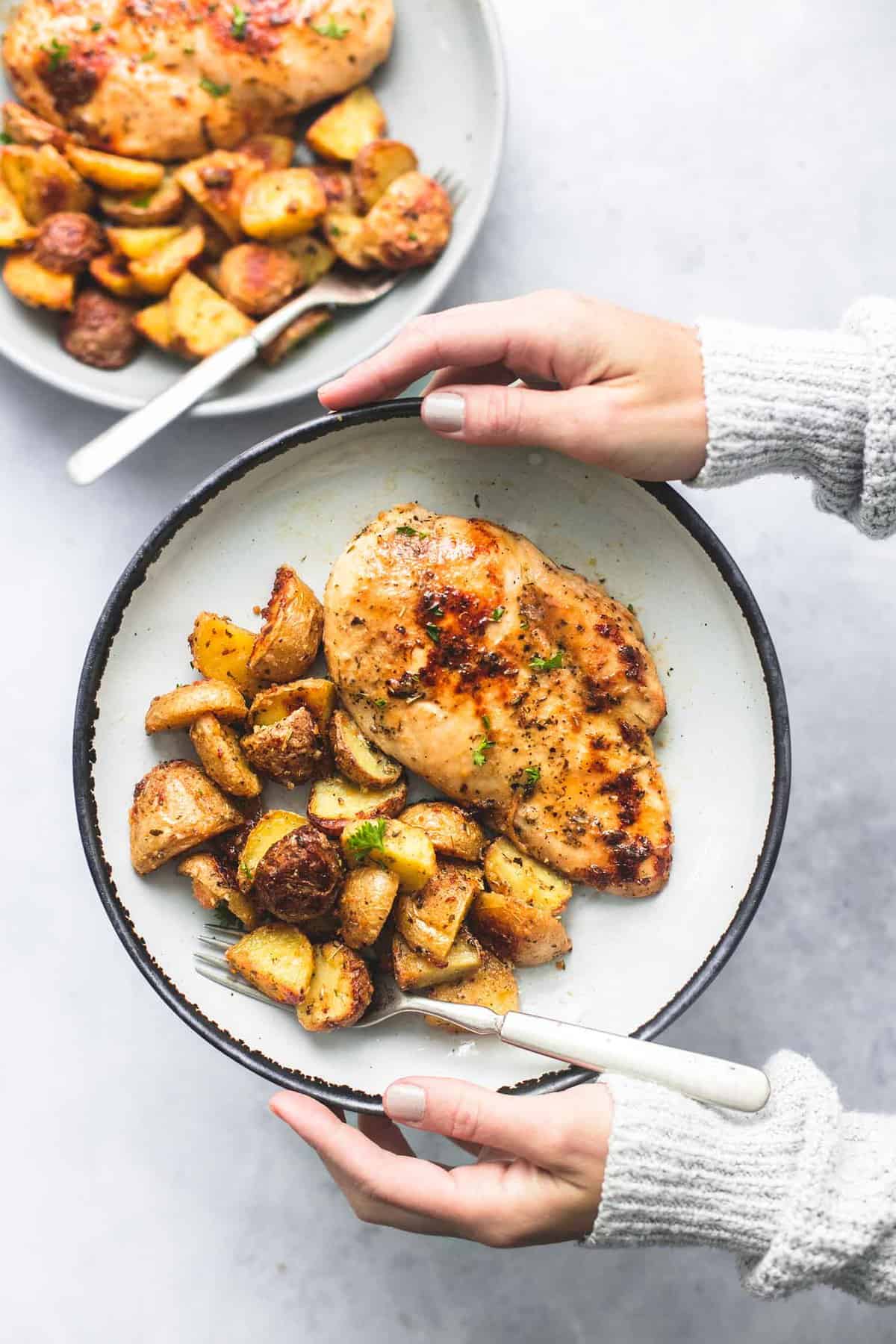 top view o f hands holding a plate with a fork and sheet pan chicken and potatoes on it with another plate on the side.