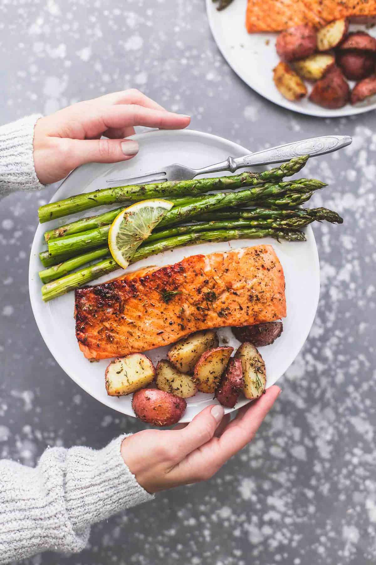 top view of hands holding a plate with a fork and sheet pan baked salmon and asparagus with potatoes on it with another plate on the side.