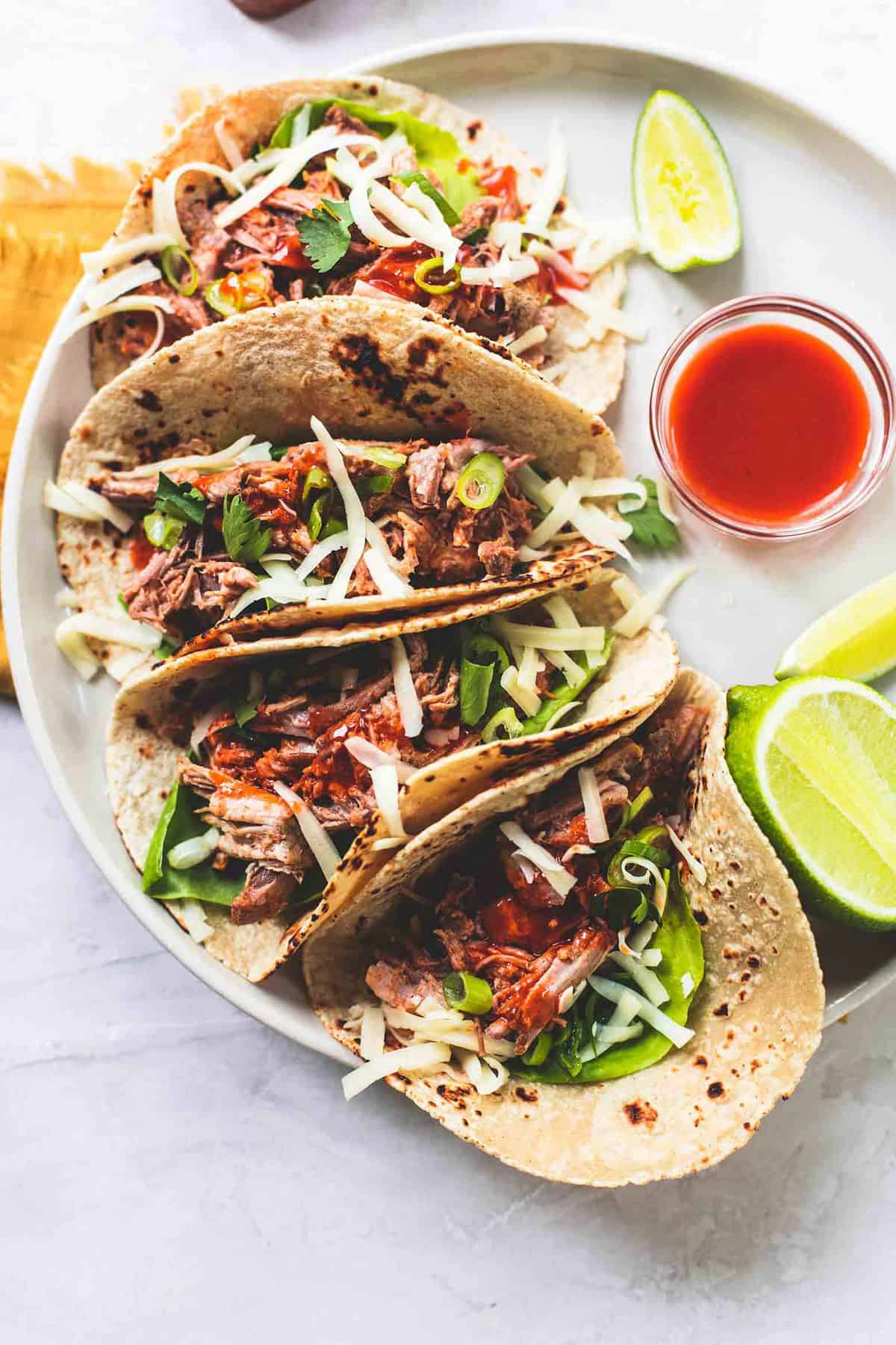 top view of slow cooker pork tacos with lime slices and a lime half and a bowl of sauce on the side all on a plate.