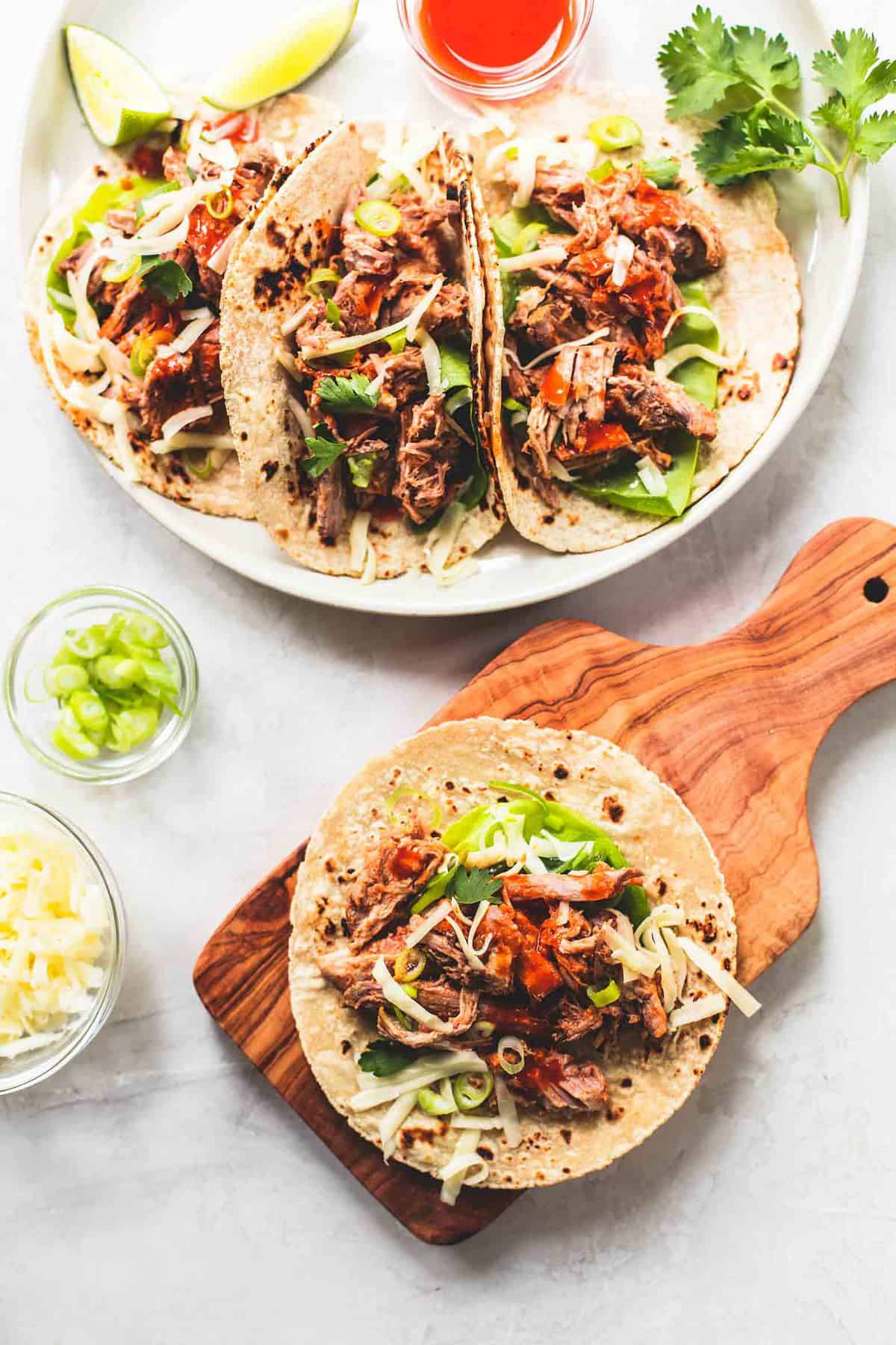 top view of a slow cooker pork taco on a wooden cutting board with bowls of toppings on the side next to a plate of tacos with sauce on the side.