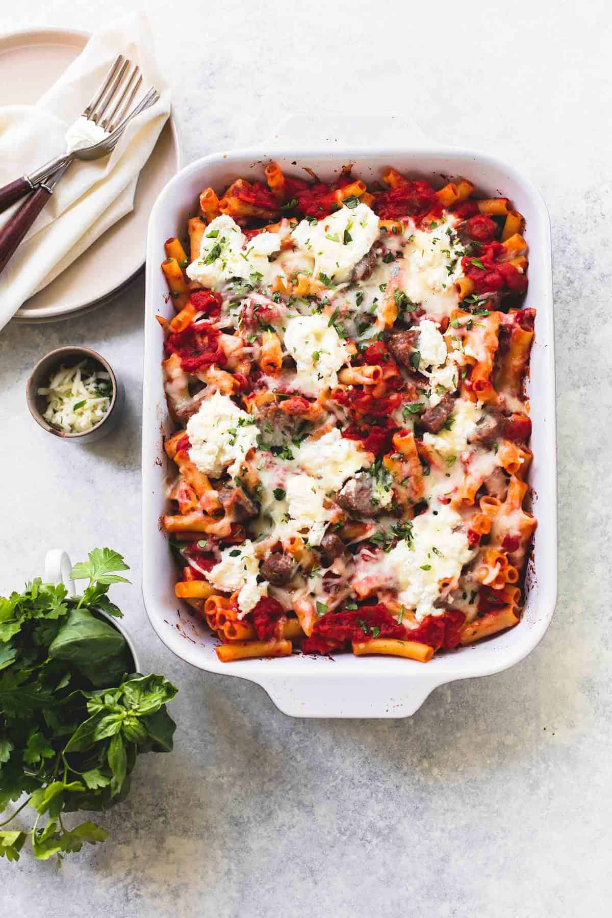 top view of baked ziti with ricotta and sausage in a baking pan with a plate with a cloth napkin and forks on top, a small bowl of mozzarella cheese, and parsley leaves on the side.