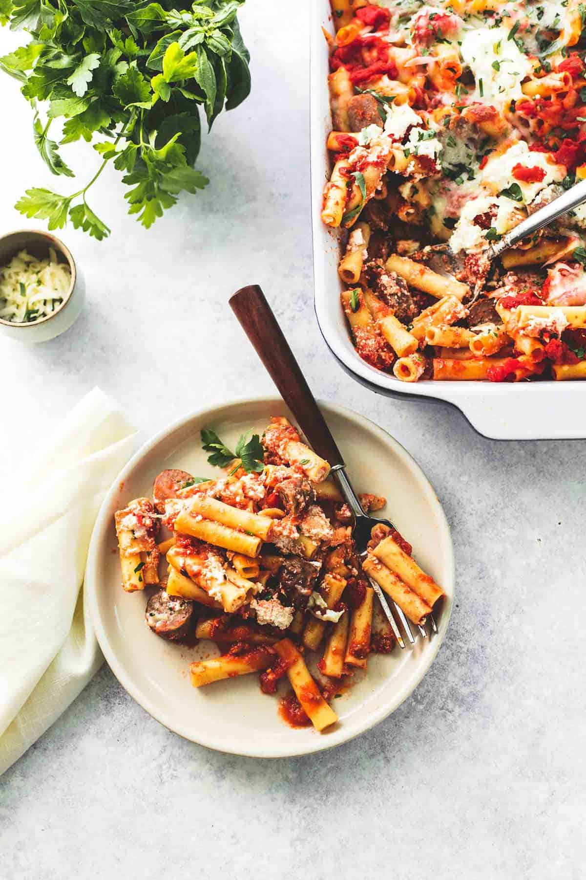 top view of baked ziti with ricotta and sausage with a fork on a plate with more ziti in a baking pan, mozzarella cheese in a small bowl and parsley leaves all in the background.