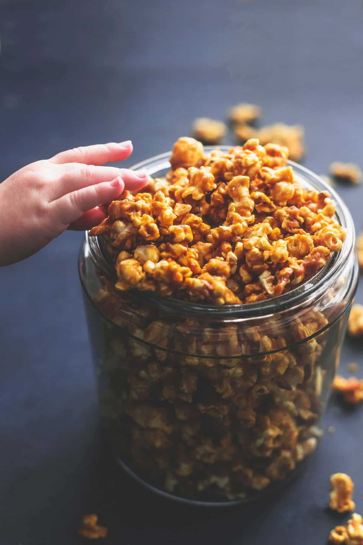 a child's hand grabbing some caramel corn from a tall glass jar.