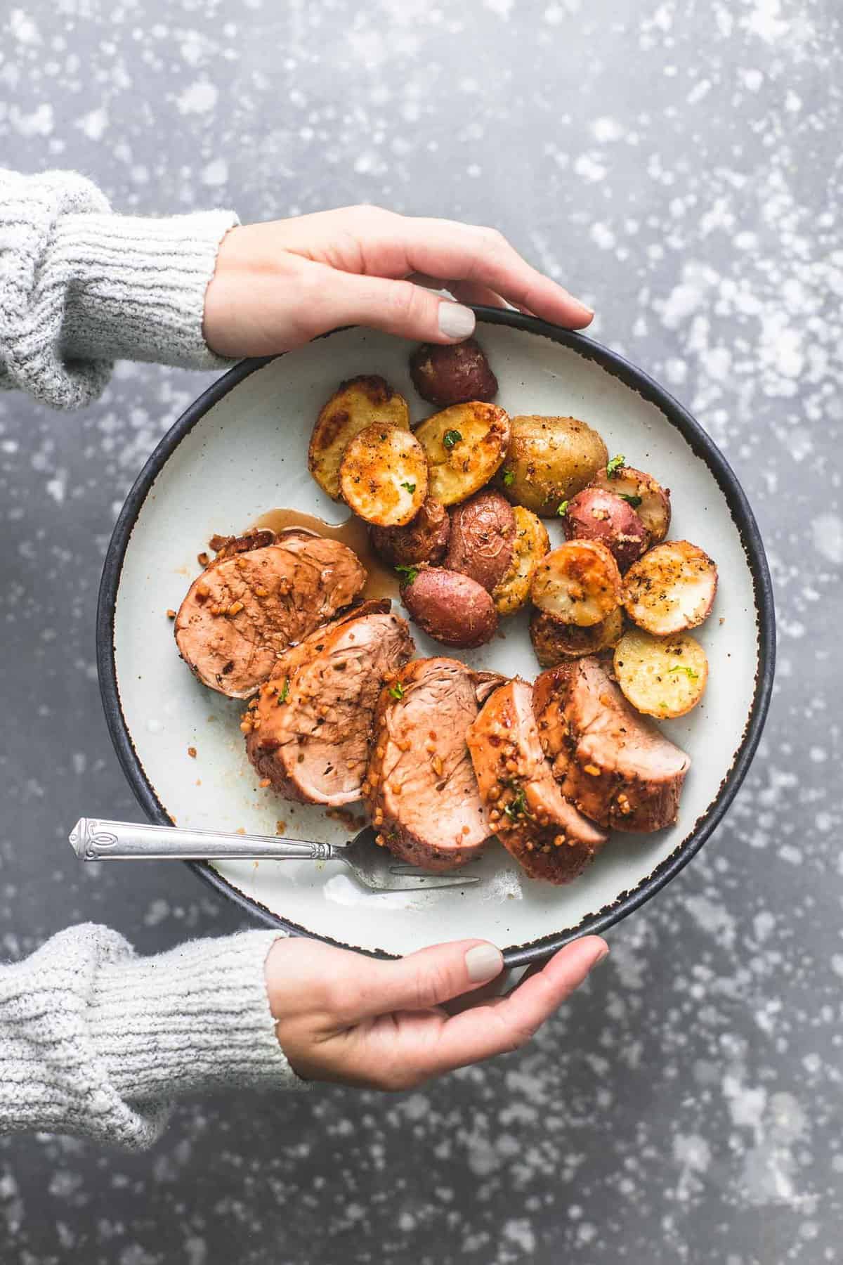top view of hands holding a plate with a fork and sheet pan pork tenderloin and potatoes on it.