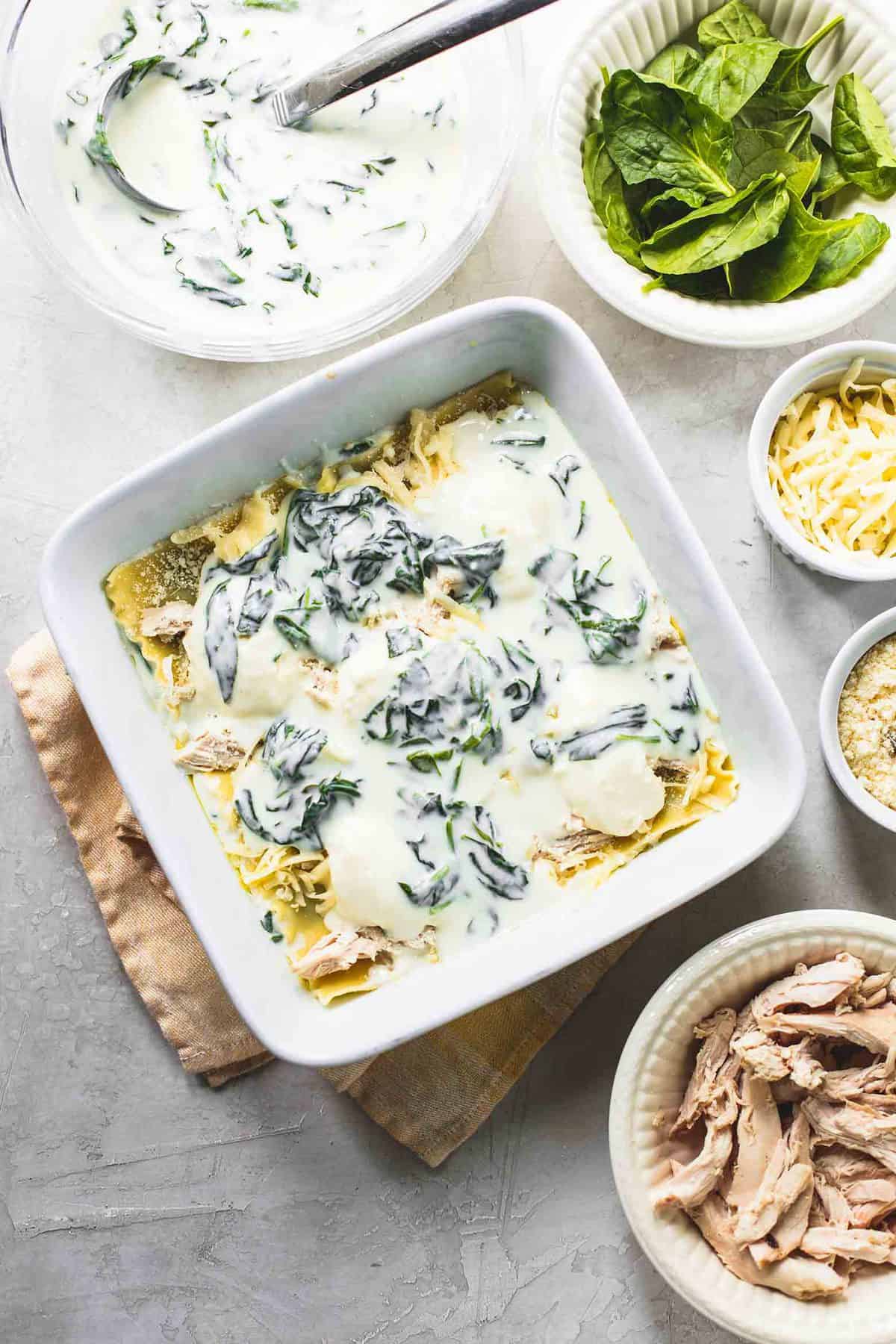 top view of white chicken lasagna with spinach in a baking pan with bowls of each ingredient on the side.