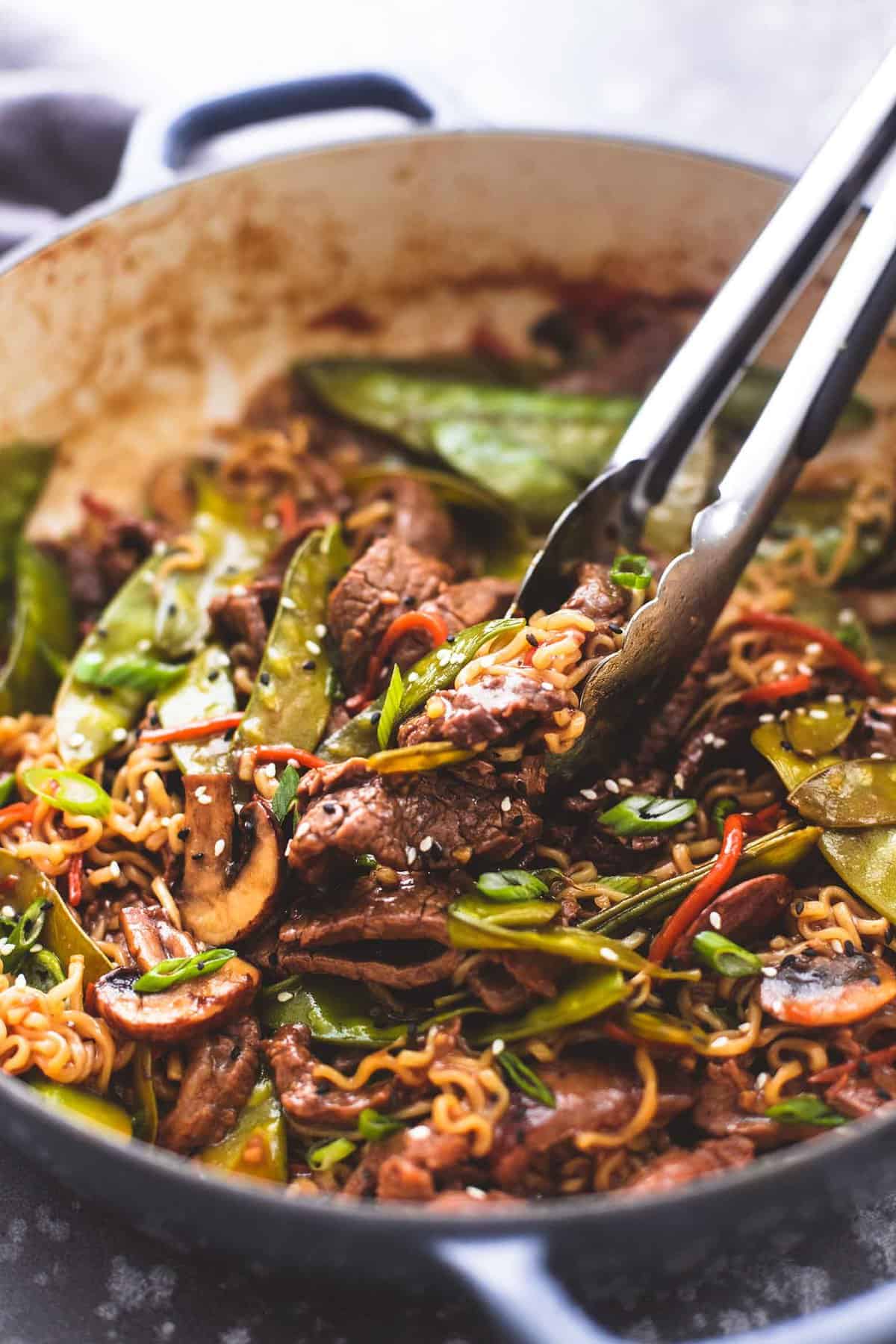 tongs lifting up some beef noodle stir fry from a pan.