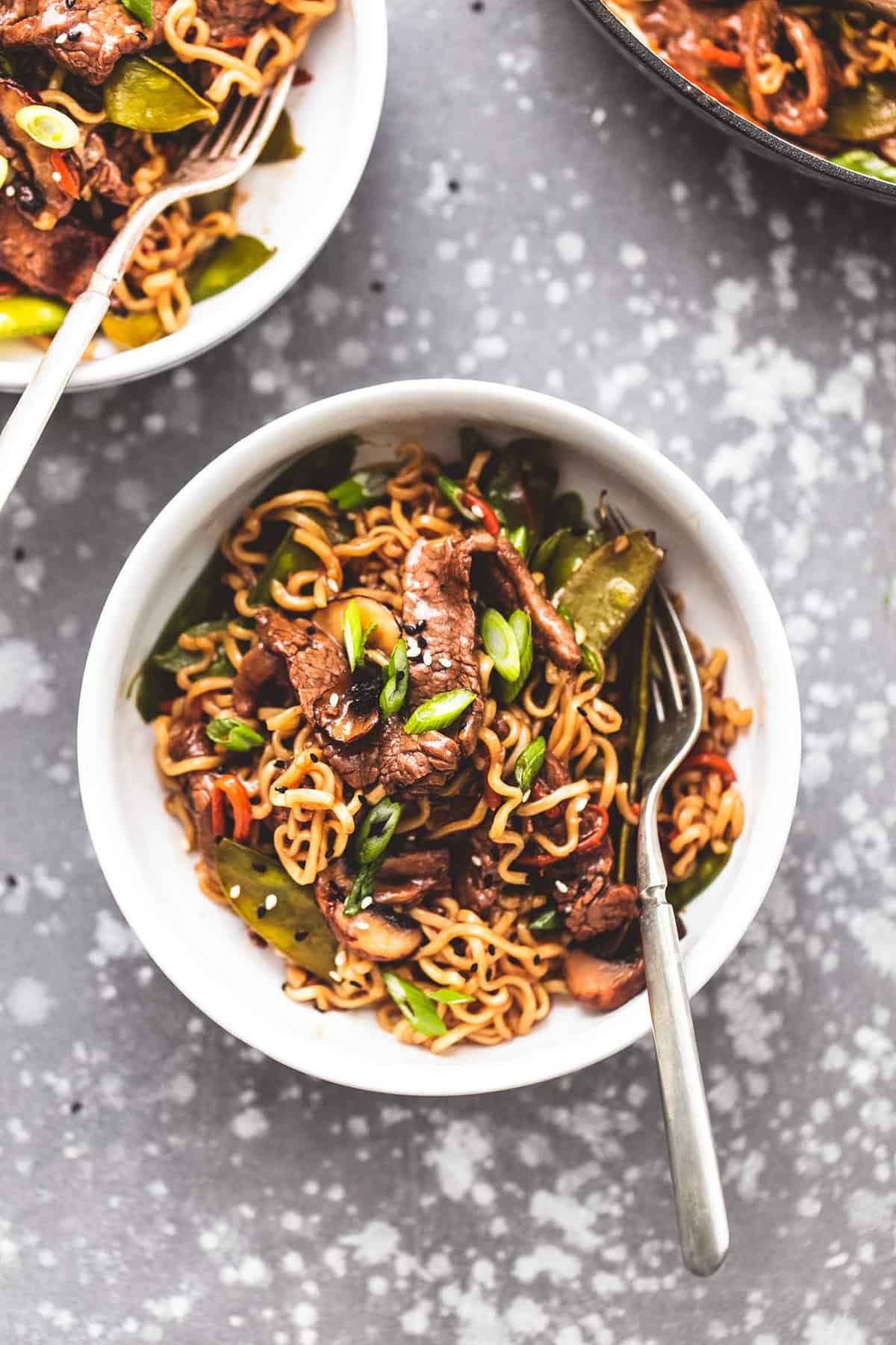top view of beef noodle stir fry with a fork in a bowl with another bowl and a pan of stir fry on the side.