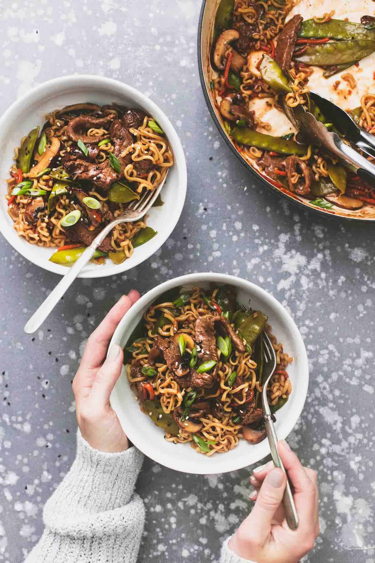 top view of a hand holding the side of a bowl with beef noodle stir fry in it and holding a fork with the other hand with another bowl and pan of stir fry on the side.