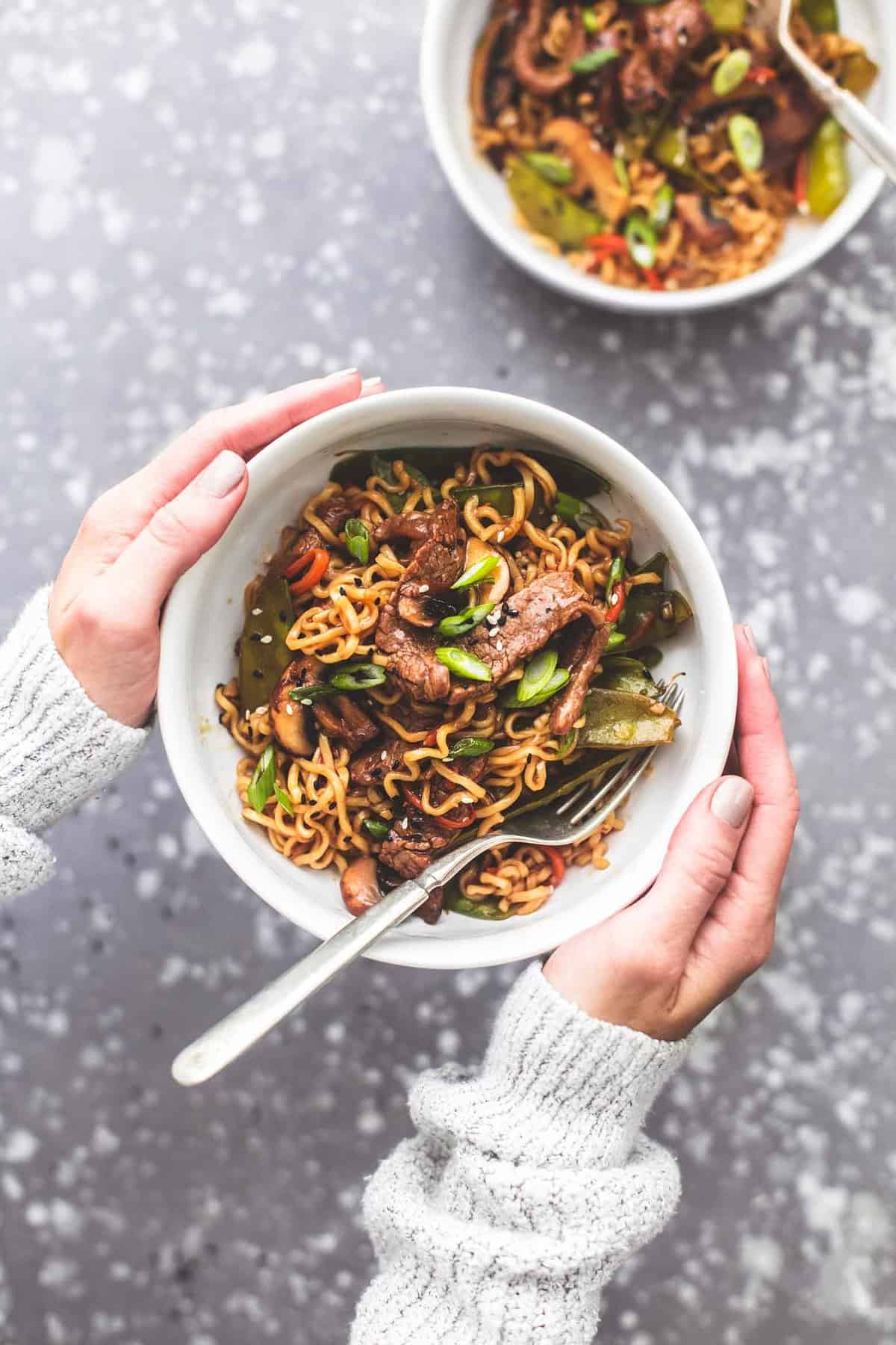 top view of hands holding a bowl of beef noodle stir fry with a fork with another bowl on the side.