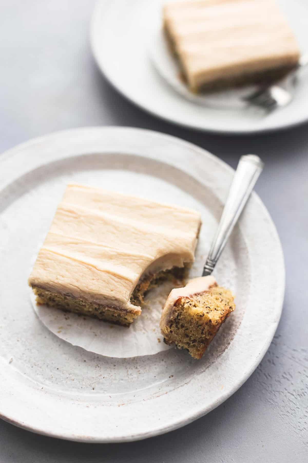 a banana bars with cream cheese frosting with a bite on a fork on the side all on a plate with another bar and fork on a plate in the background.