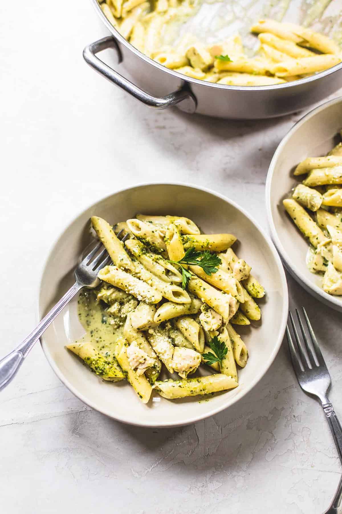 top view of creamy pesto chicken pasta with a fork in a bowl with another bowl and a pot of more pasta on the side.