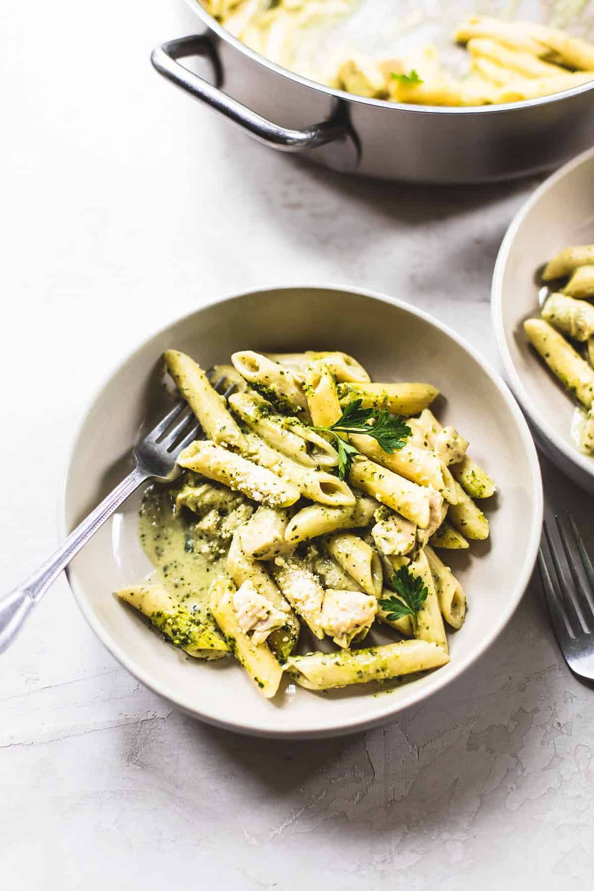 creamy pesto chicken pasta with a fork in a bowl with another fork, bowl of pasta and a pot of more pasta on the side.
