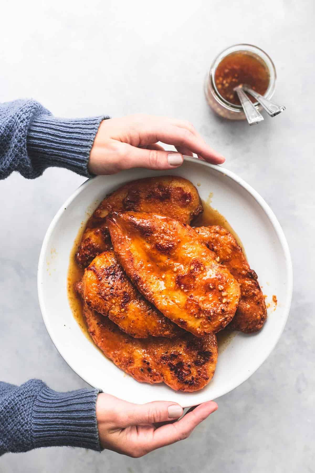 hands holding up a plate of crispy honey garlic chicken with a glass jar of sauce with spoons in the side.
