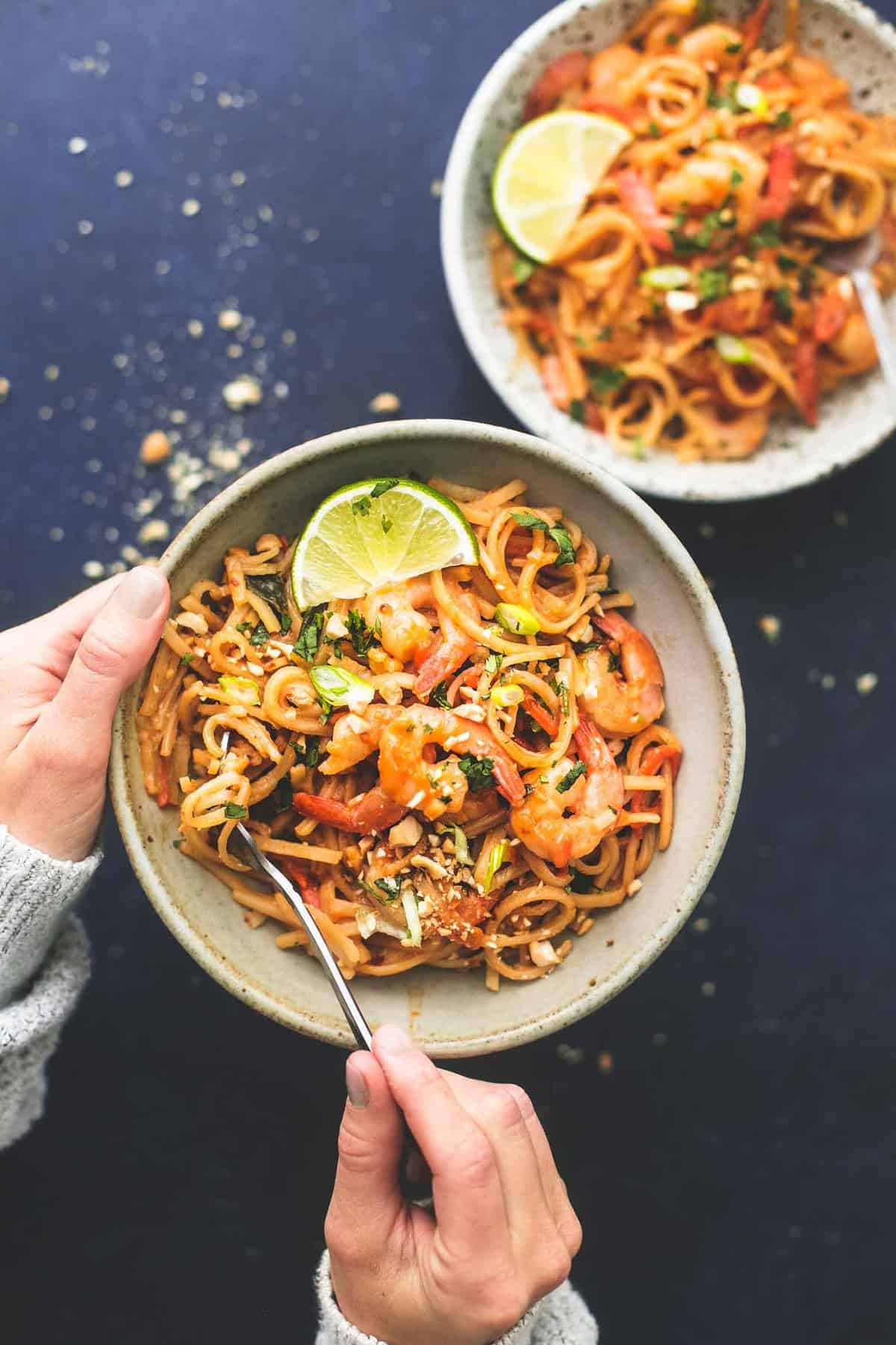 top view of a hand holding a bowl of shrimp pad Thai with peanut sauce with another hand grabbing a bite with a fork with another bowl on the side.