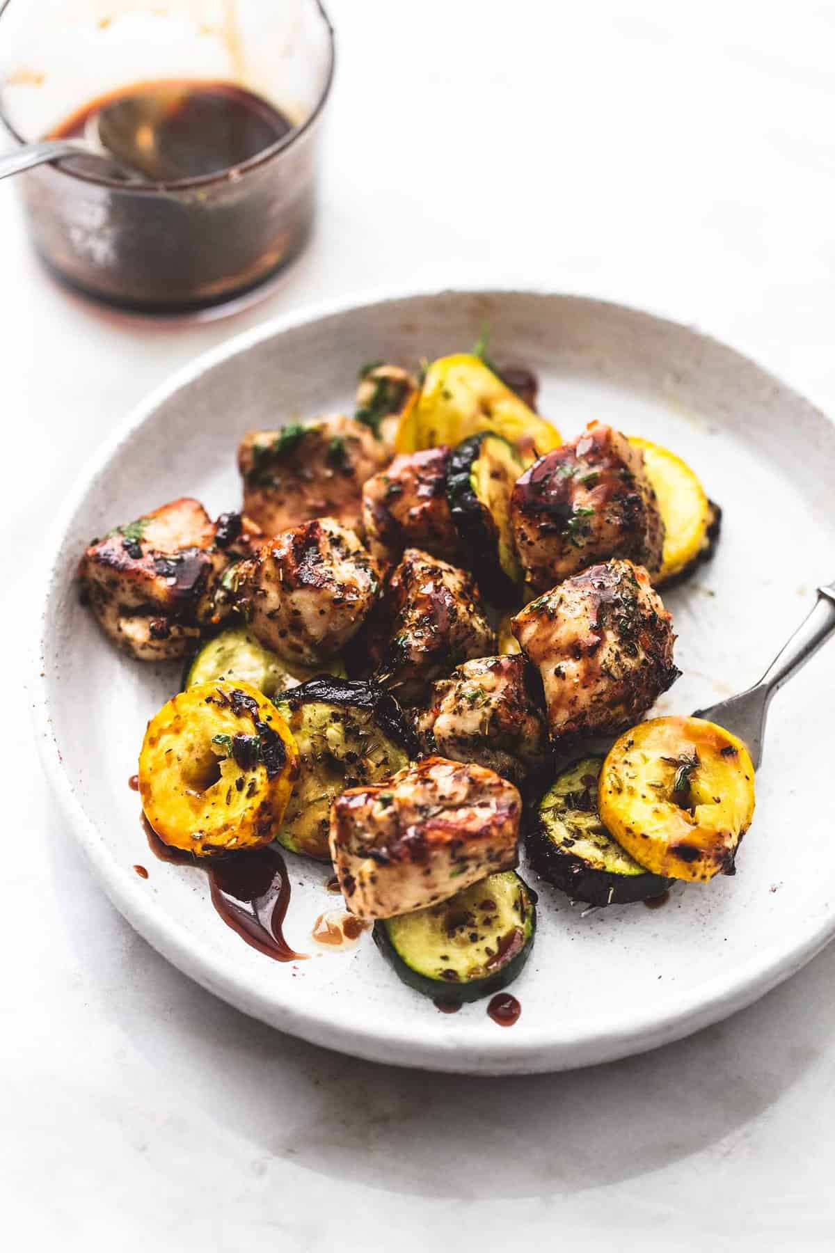 deconstructed balsamic chicken and veggie kabobs with a bite on a fork on a plate with a glass of balsamic marinade on the side.